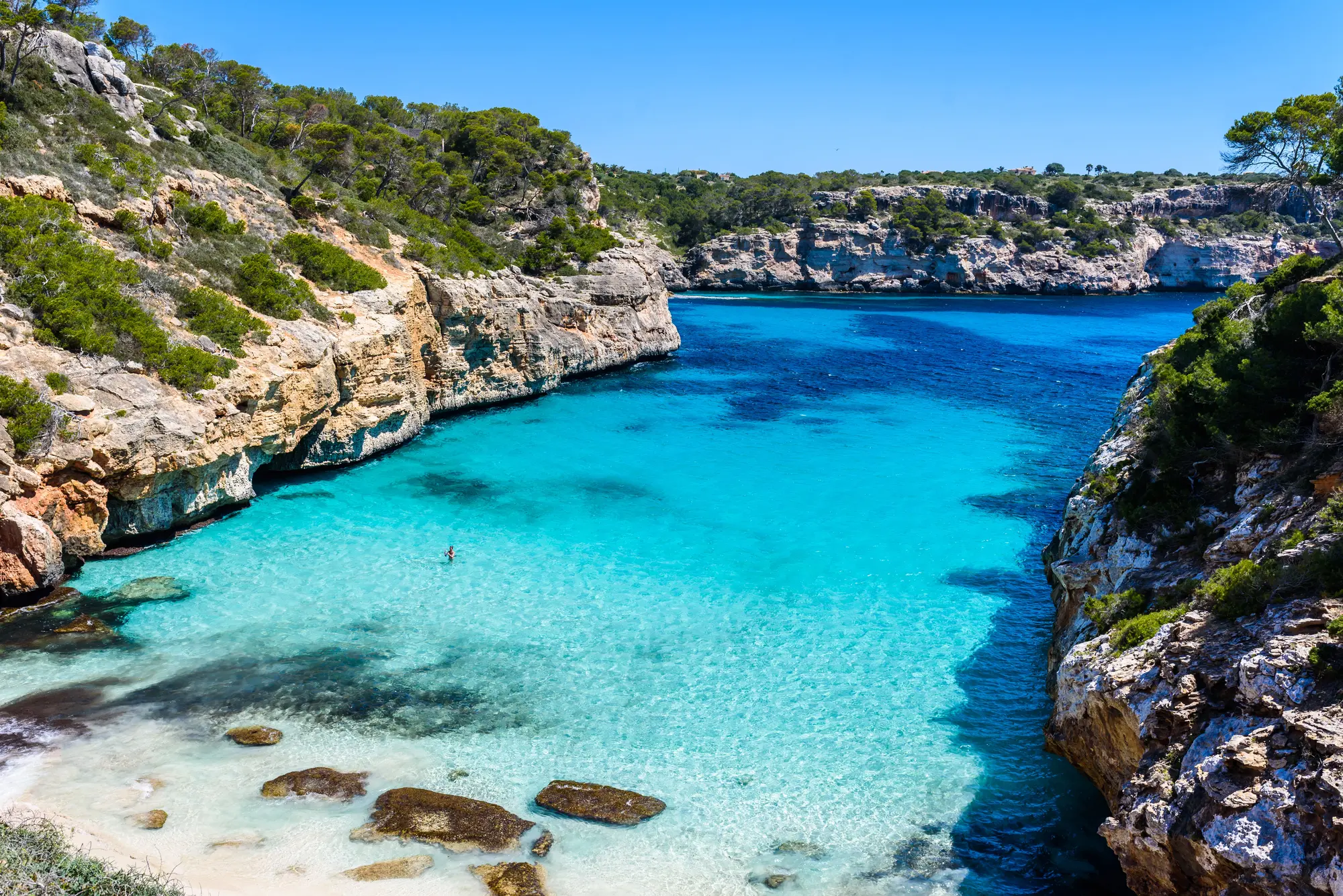 View from above of the tiny turquoise Calo des Moro with white sand and stones in the water, surrounded by rugged cliffs on a boat tour from Cala Figuera.