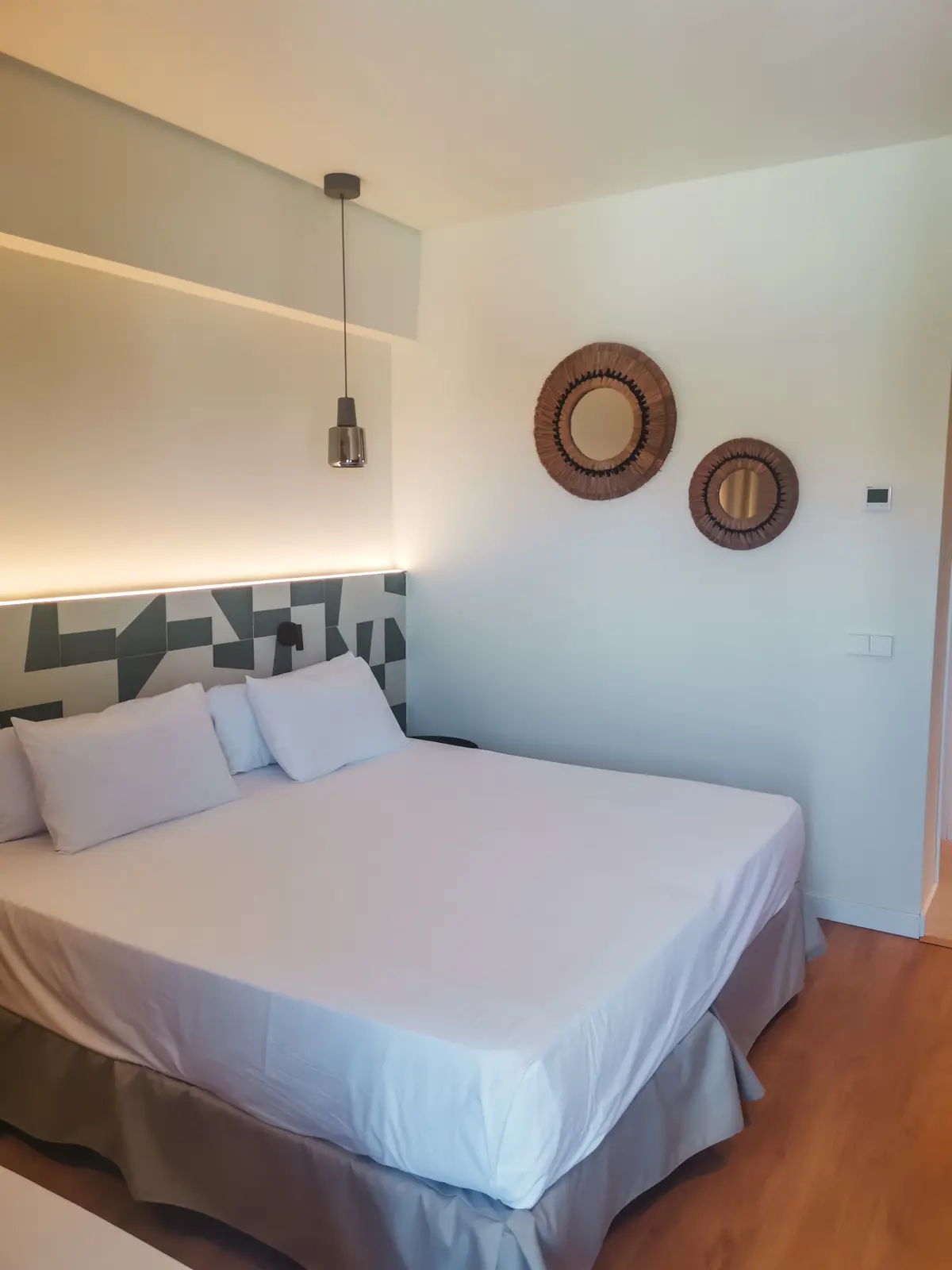 Double bed with white sheets with a small lamp hanging above it, straw mirrors on the wall and white and green tiles in a random pattern on the headboard at Hotel Santanyi Port, Cala Figuera.