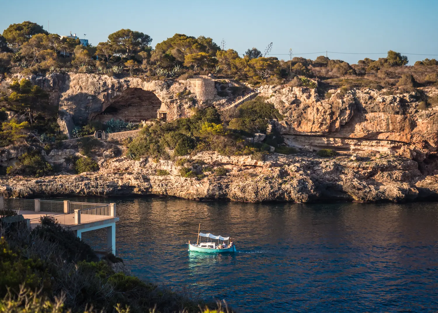 Small turquoise sailboat on the dark blue water in Cala Figuera with a cliff covered in trees in the background.