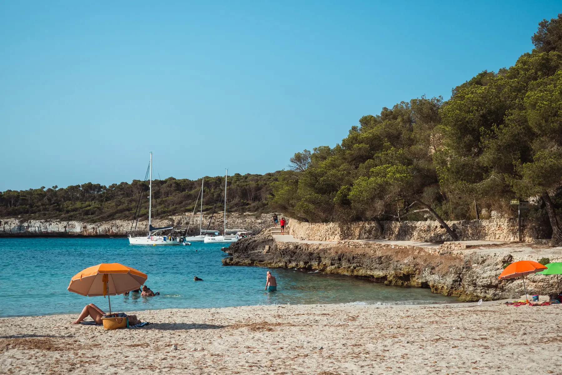 Woman on Cala Mondrago beach with an orange umbrella and orange bag with turquoise water and two sailboats in the background, in Mondrago Natural Park, Mallorca.