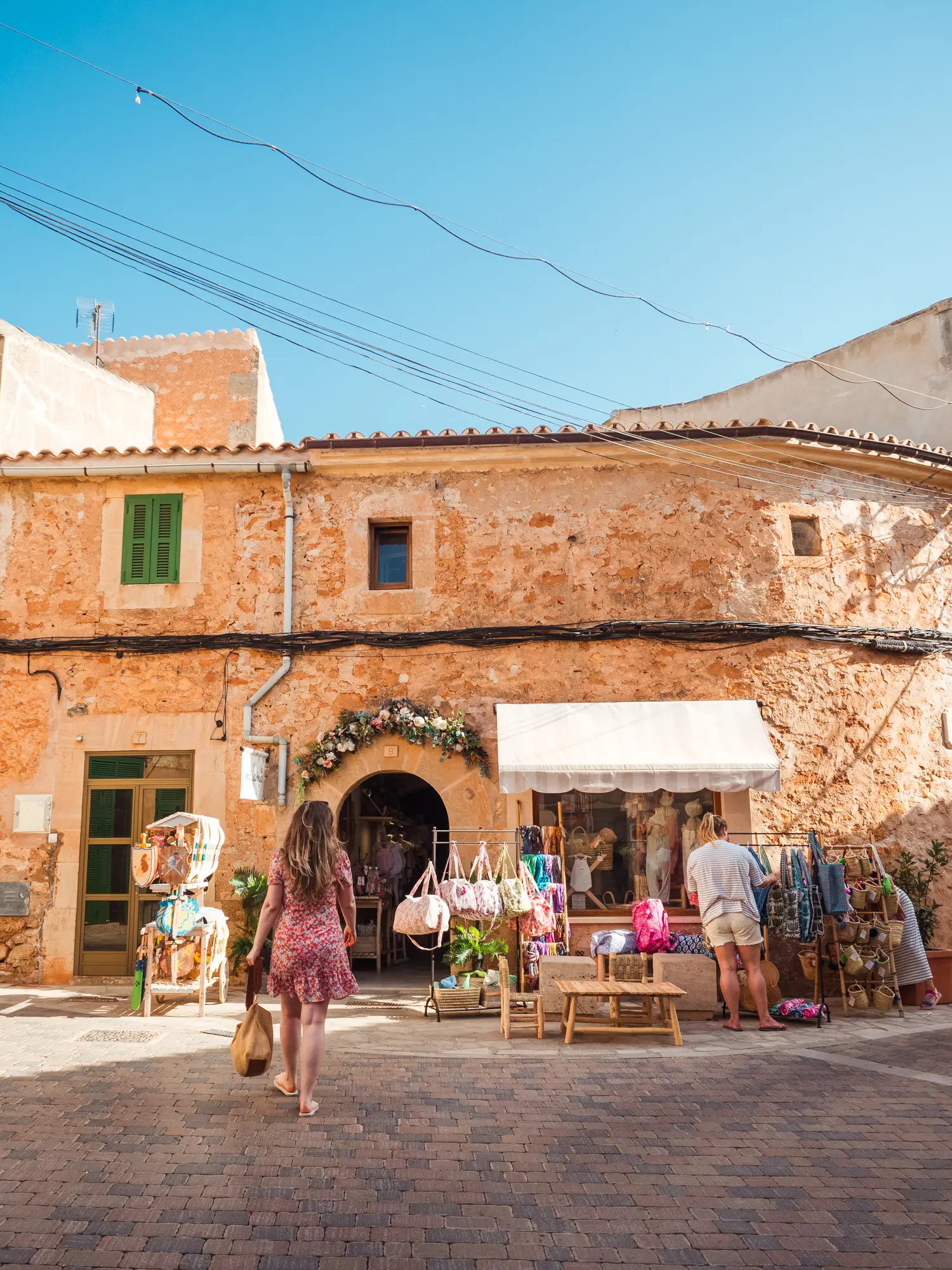 Woman with long hair, wearing a pink dress, walking up to a little shop in a sandstone building at Santanyi Market in Mallorca.