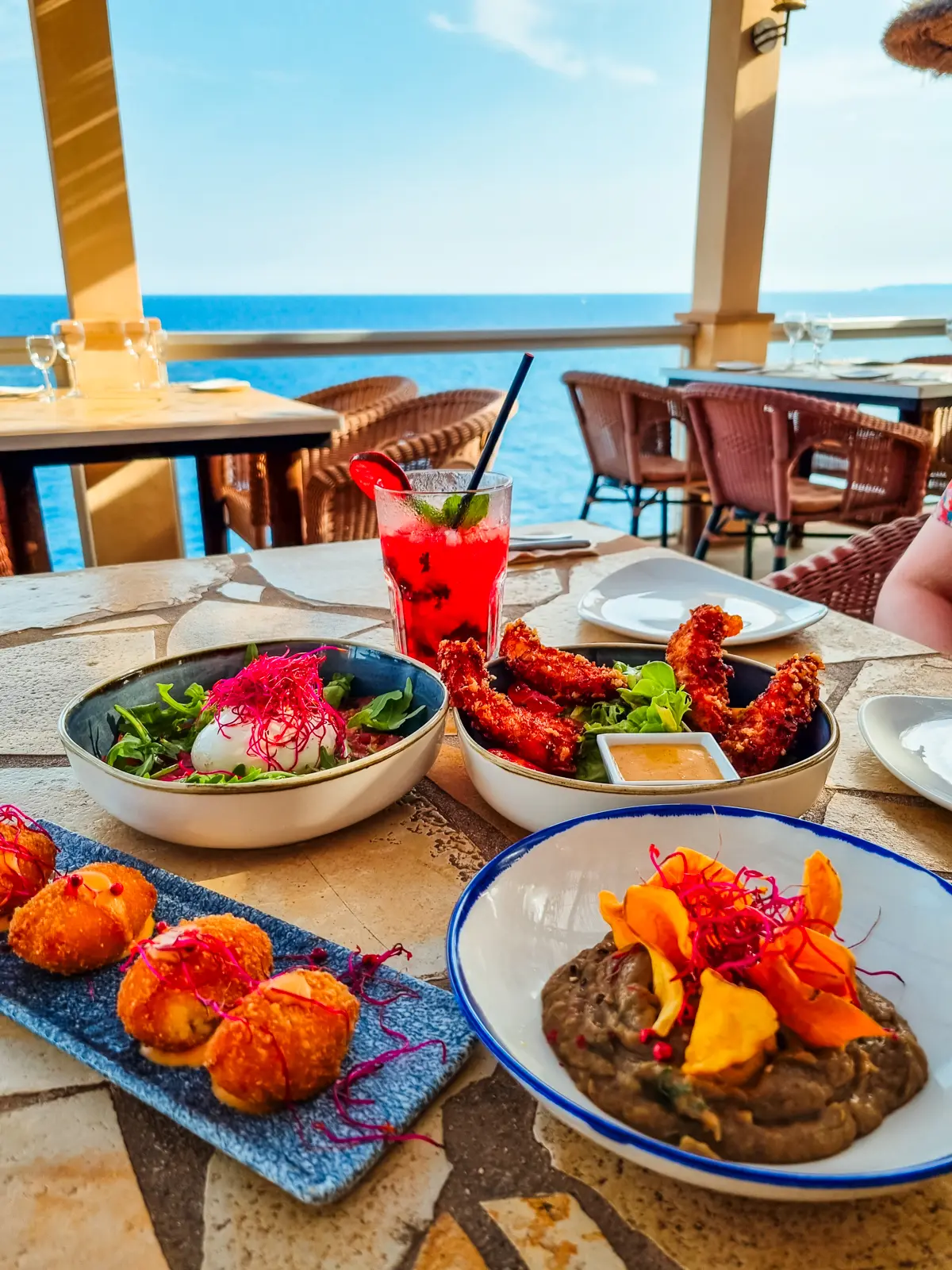 Three white bowls and a rectangle shaped blue tray with colorful tapas, with the ocean in the background at Pura Vida Restaurant in Cala Figuera, Mallorca.