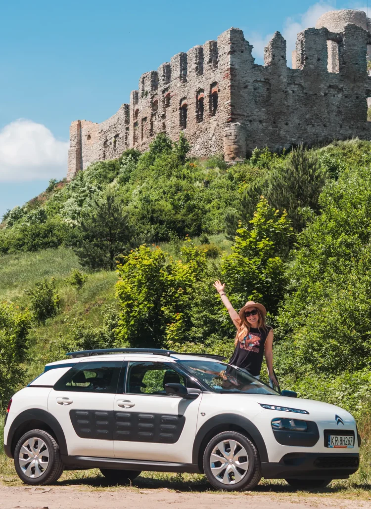 Woman wearing a black top and beige har, reaching out the window of a white and black Citroën C4 Cactus, with one hand in the air pointing toward a castle ruin on a hill, after renting a car in Krakow.