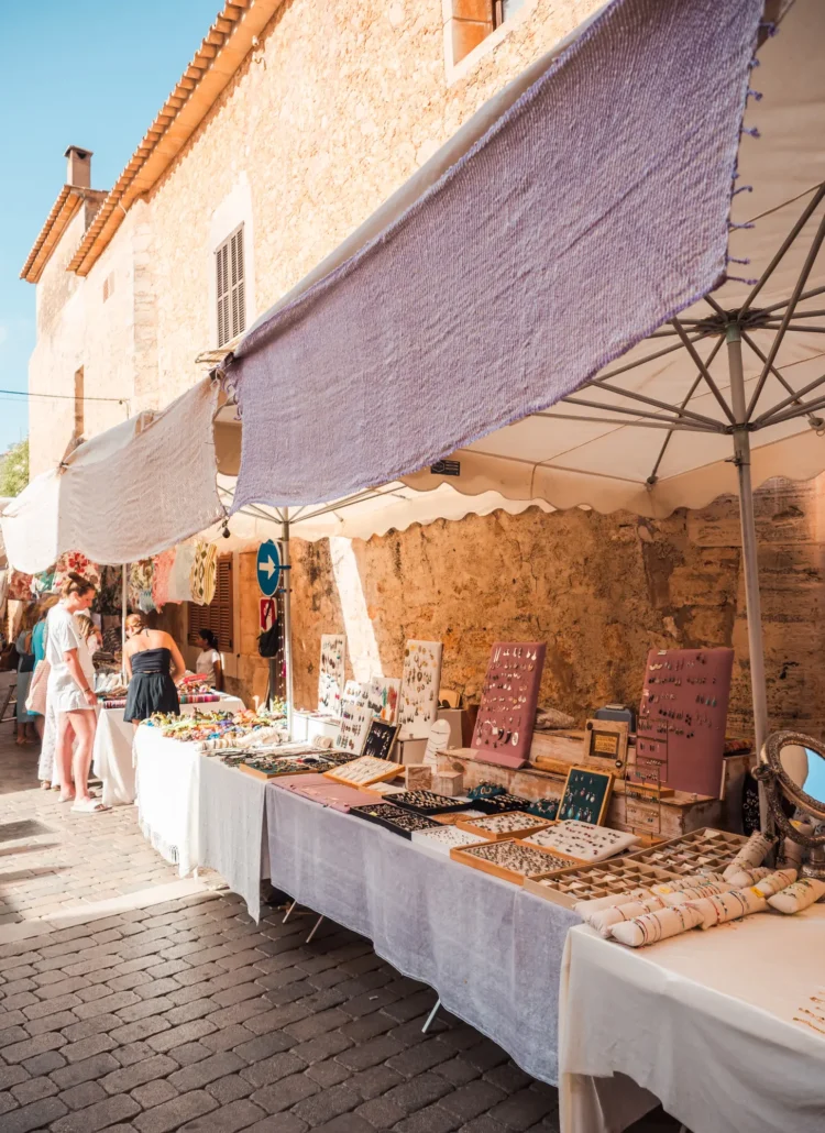 Table with white table cloths displaying jewelry under parasols against a sandstone building at Santanyi Market in Mallorca.