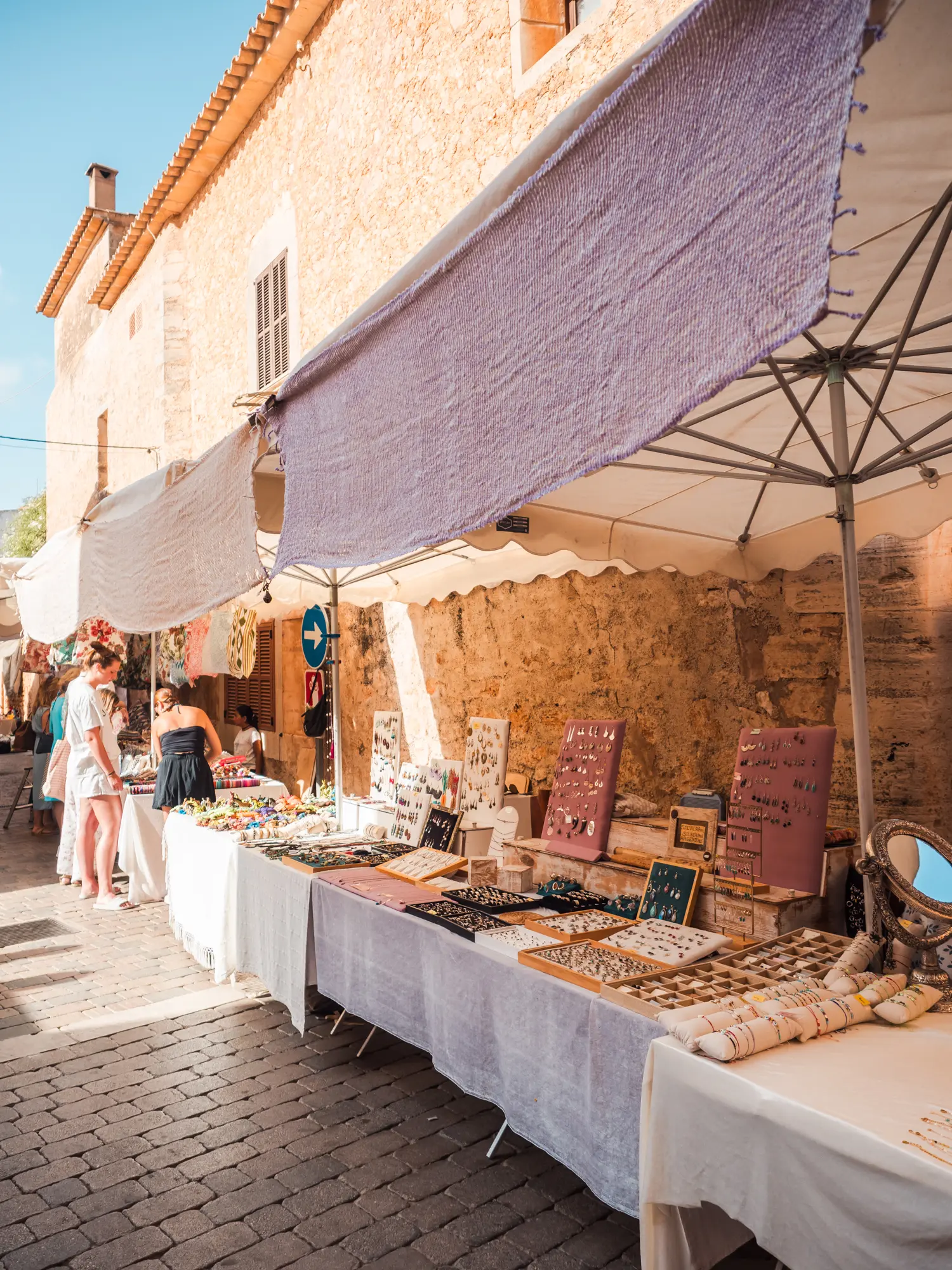 Table with white table cloths displaying jewelry under parasols against a sandstone building at Santanyi Market in Mallorca.