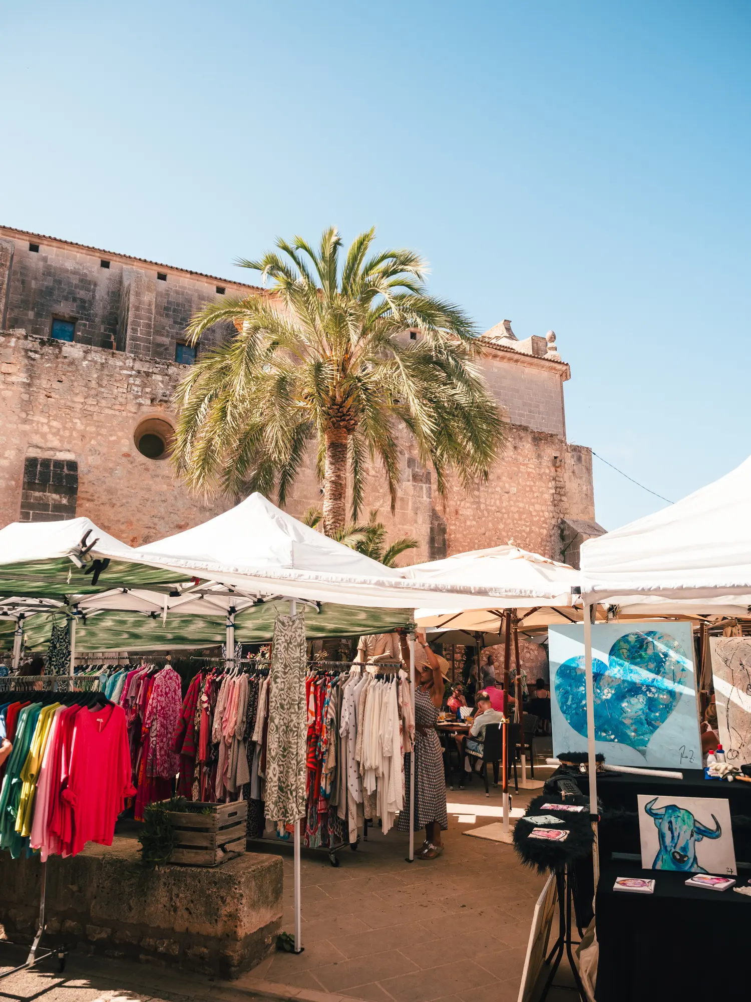 Large sandstone building with a tall palm tree and white tents with clothes in front at Santanyi Market in Mallorca.