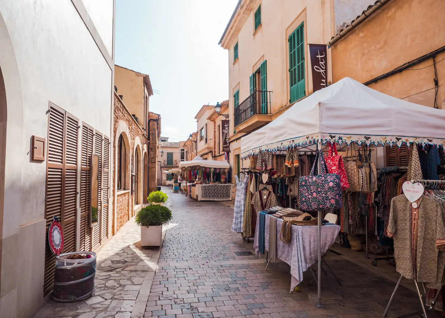 Narrow cobbled road between sandstone houses with green shutters and a stall selling clothes and bags at Santanyi Saturday Market.