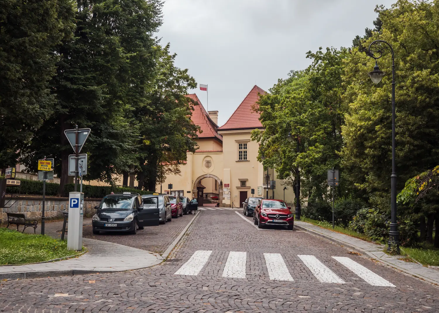 Cobbled street with green trees on both sides and a taxi queue, leading up to a light yellow house outside Wieliczka Salt Mine in Krakow.