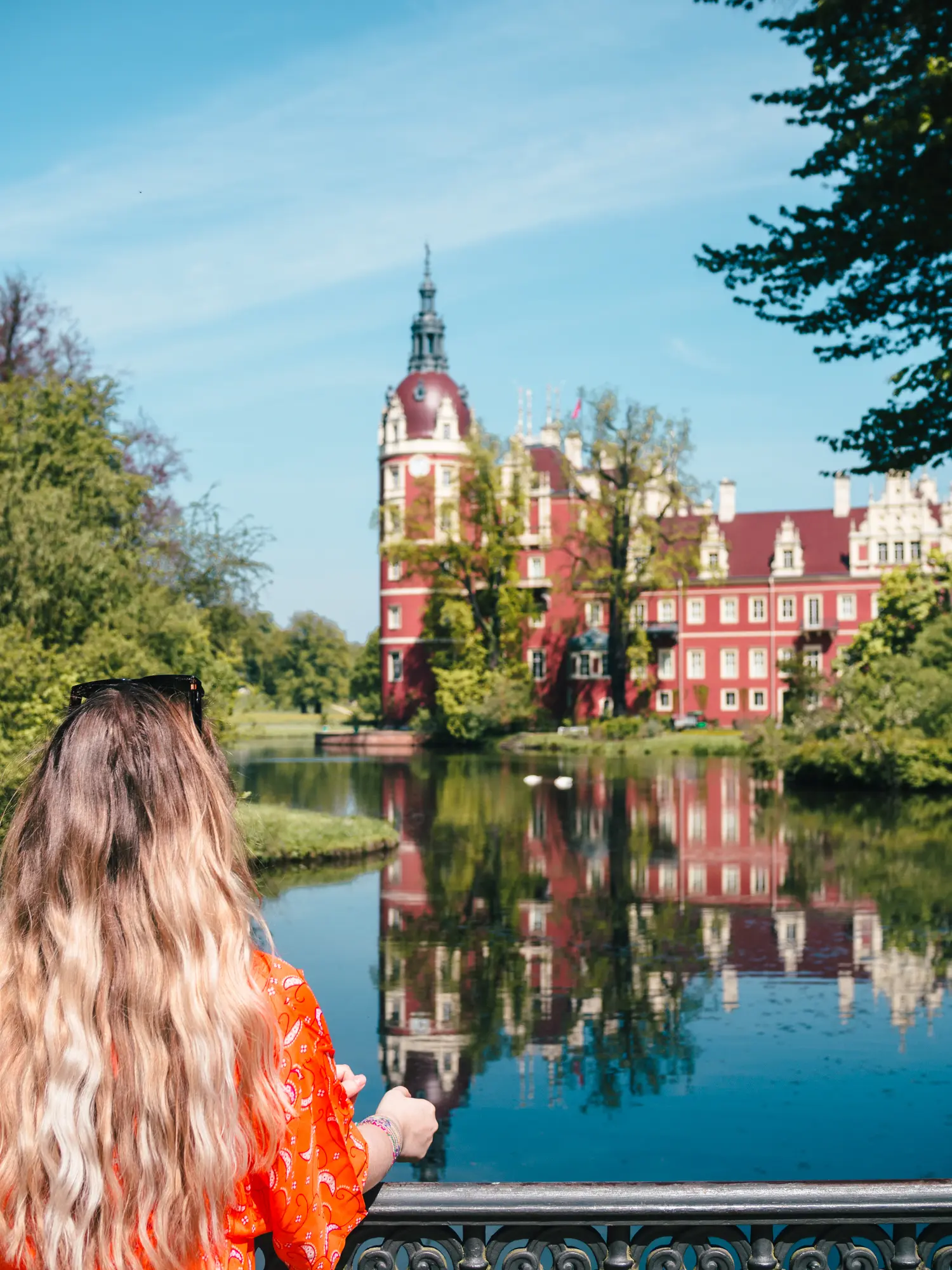 Woman with long dark blonde hair, wearing an orange top, looking out over the lake in front of the red Schloss Muskau Castle on a road trip from Krakow. 