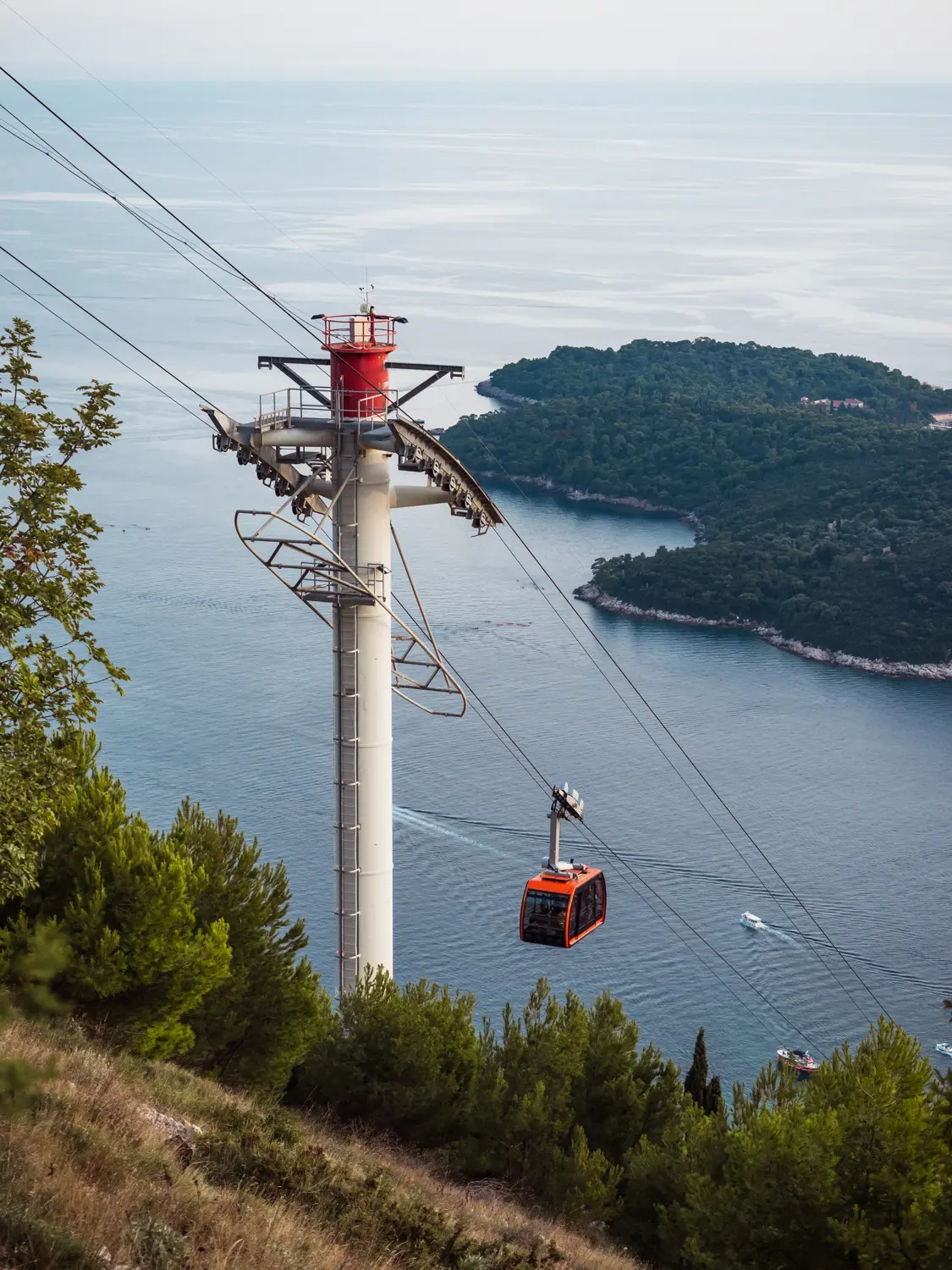 Red cable car going up to Mount Srs viewpoint in Dubrovnik, with Lokrum Island and the ocean in the background.