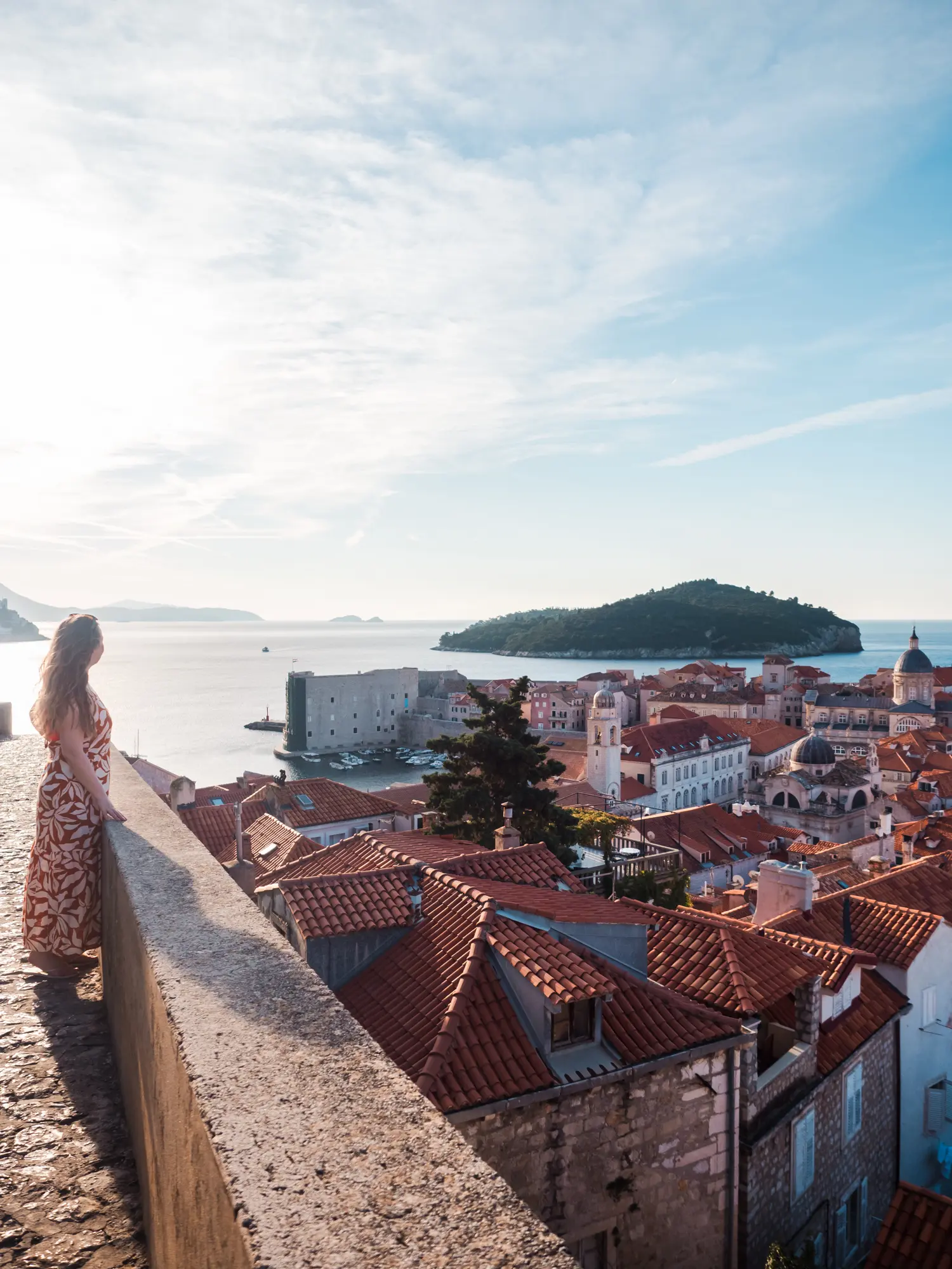 Woman wearing a long white and orange dress standing on the old stone City Wall of Dubrovnik looking out over the Old Town on a sunny morning, included in the Dubrovnik Pass.