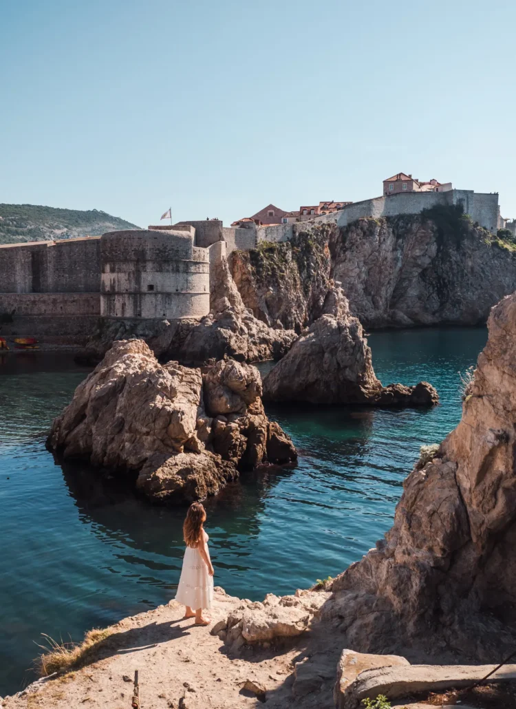 Woman with long hair wearing a white dress standing on a stone by the blue ocean looking over at the old City Wall, included in the Dubrovnik Pass.