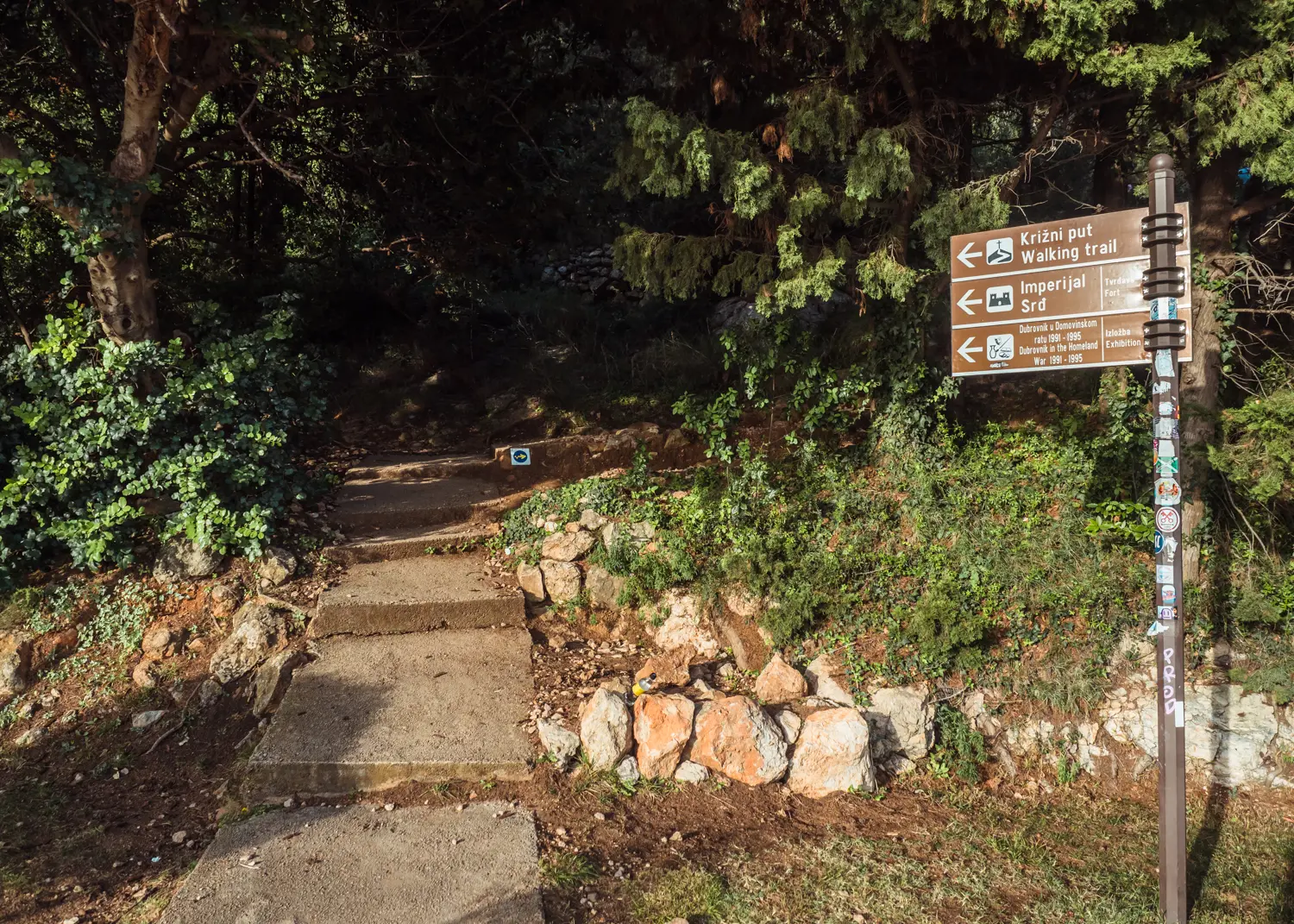 Three brown signs to the right, paved path into the forest, the start of the hike to Mount Srd Dubrovnik viewpoint.