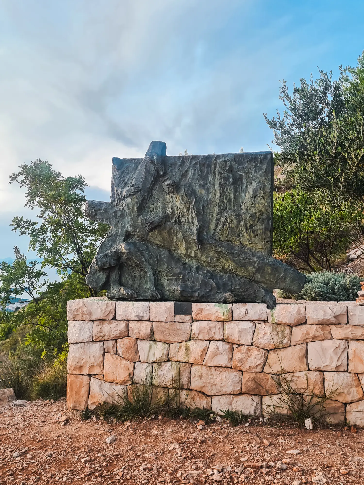 Cross metal artwork on a stone wall with trees in the background, showing the hiking route to Mount Srd, the best sunset viewpoint in Dubrovnik.