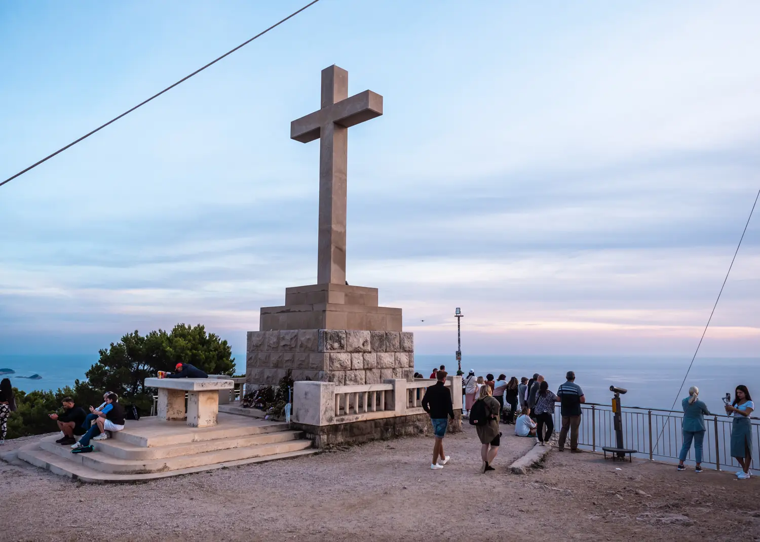 Large stone cross with people around it looking at a pastel colored sunset over Dubrovnik from Mount Srd Panorama viewpoint.