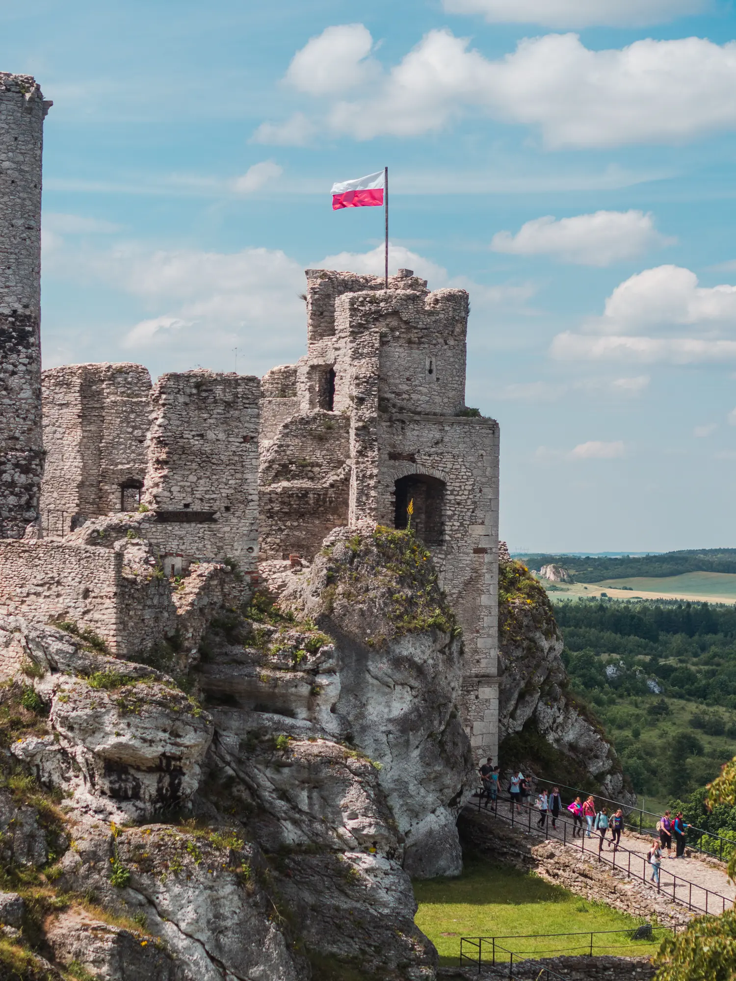 Grey stone tower at Ogrodzieniec Castle ruin with a Polish flag blowing in the wind, people walking up the entrance on a sunny day.