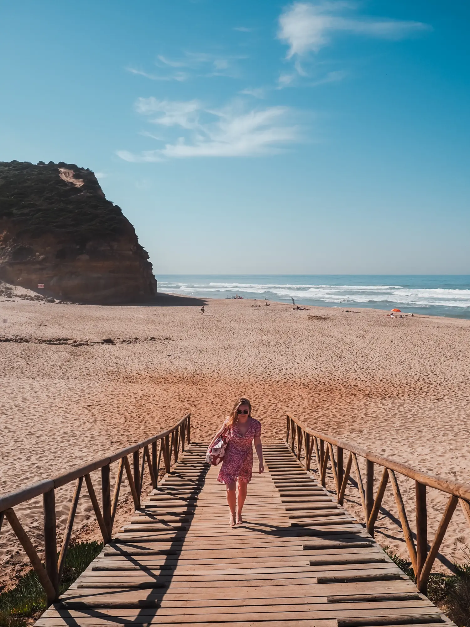 Woman in a pink dress holding a burgundy beach bag walking up a wooden path with Praia de São Julião beach and mountain in the background, surfing Ericeira.