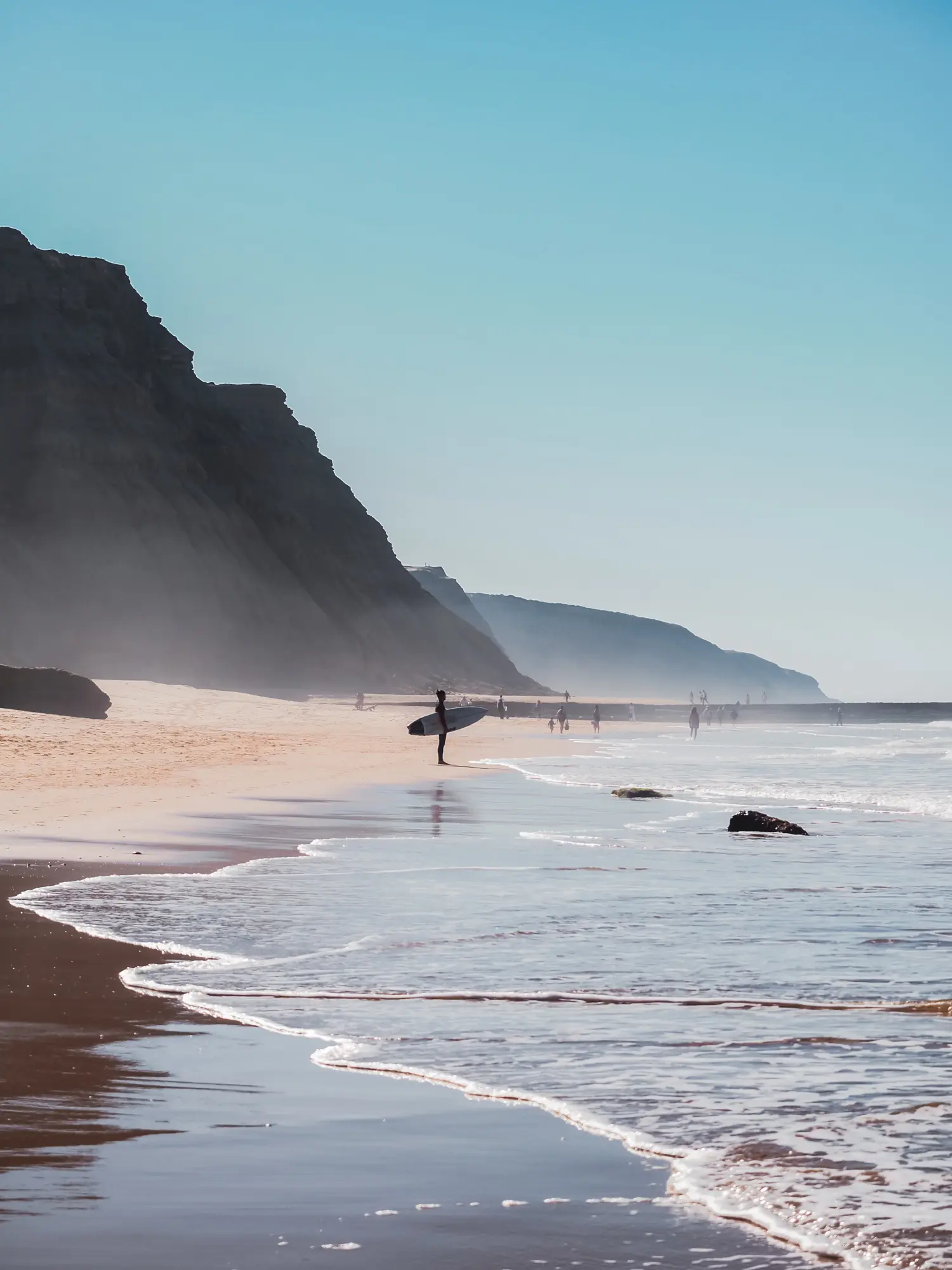 Surfer holding a surfboard standing at the shoreline in a distance on Praia do São Julião beach with mountains in the background, in Ericeira.