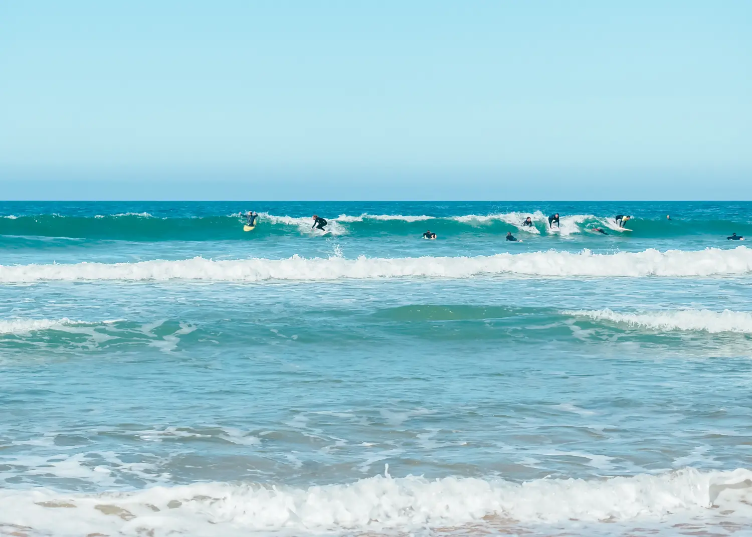 Busy line-up of surfers in wetsuits in the turquoise ocean at São Julião surfing beach on a sunny day in Ericeira.