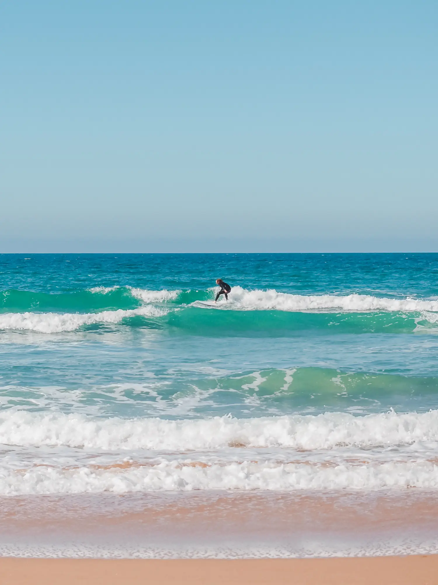 Surfer in a black wetsuit on a small wave in the turquoise ocean of Ericeira on a sunny day, Portugal's surfing city.