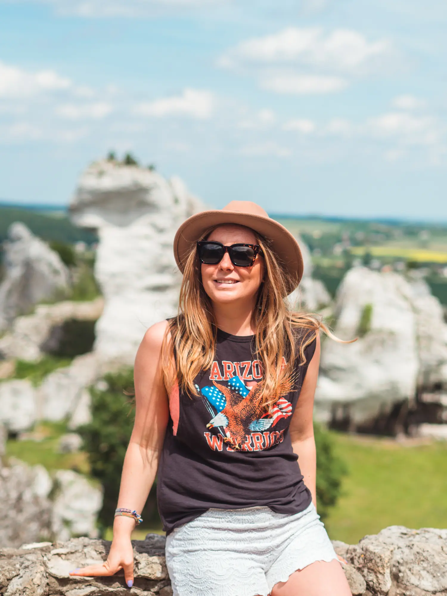 Woman with long dark blonde hair, wearing a beige hat, white shorts, sunglasses and a black tank, sitting on a stone wall in Ogrodzieniec Castle on a sunny day.