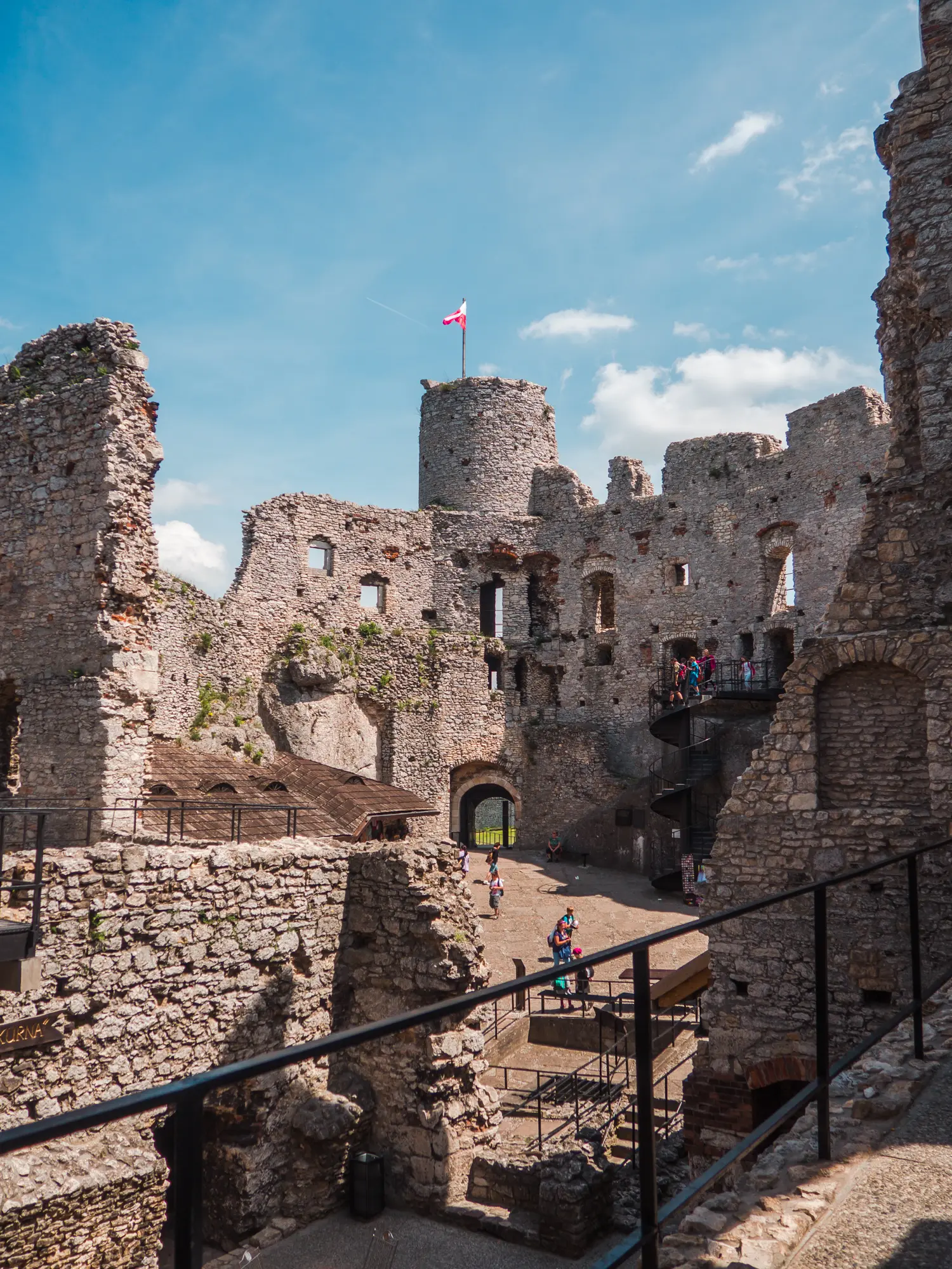 Sun shining into the haunted Ogrodzieniec Castle ruin made from grey stone, looking towards a tower with the Polish flag blowing in the wind.