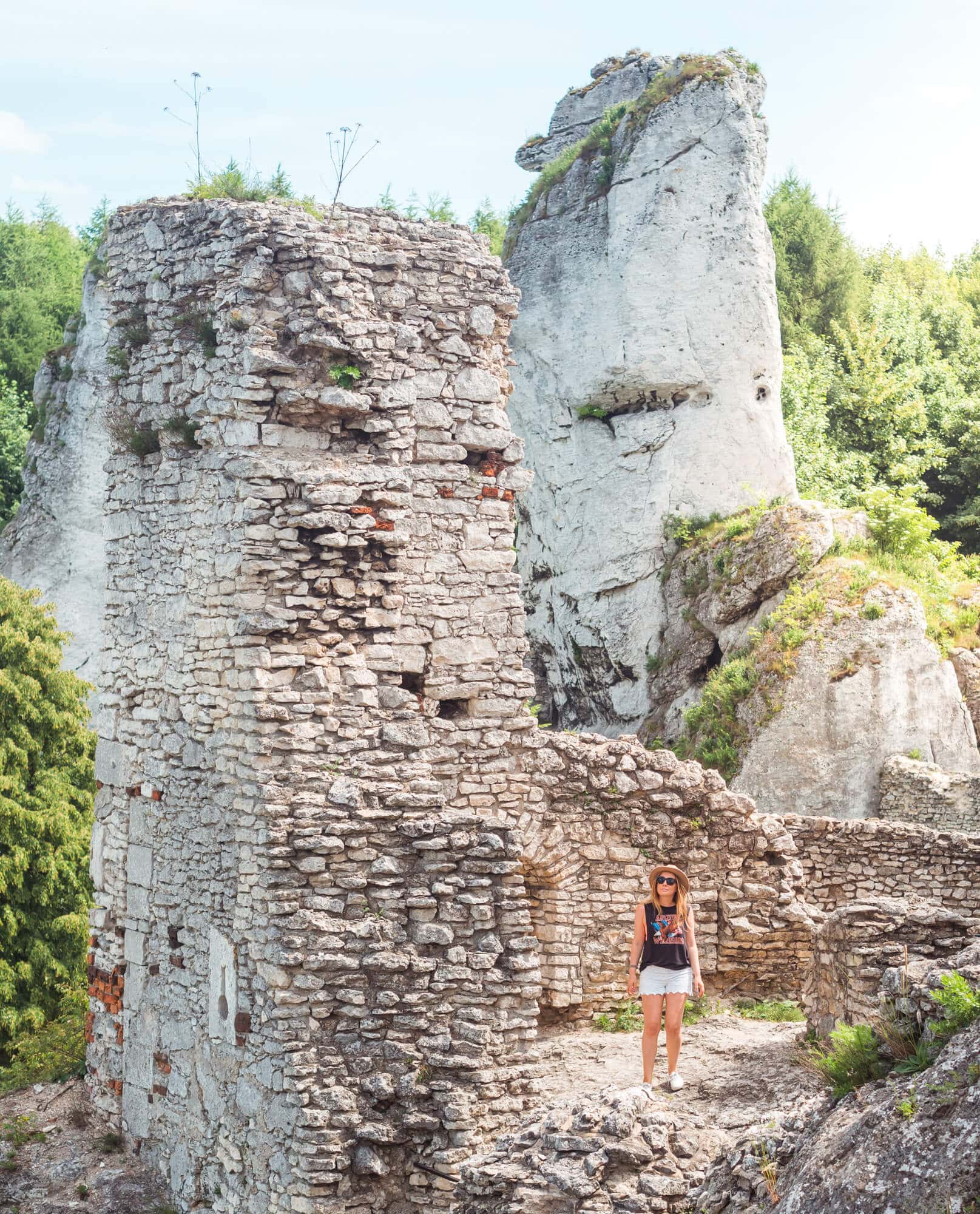 Woman wearing white shorts and a black top standing next to a stone wall ruin with a tall limestone formation in the background at Ogrodzieniec Castle.