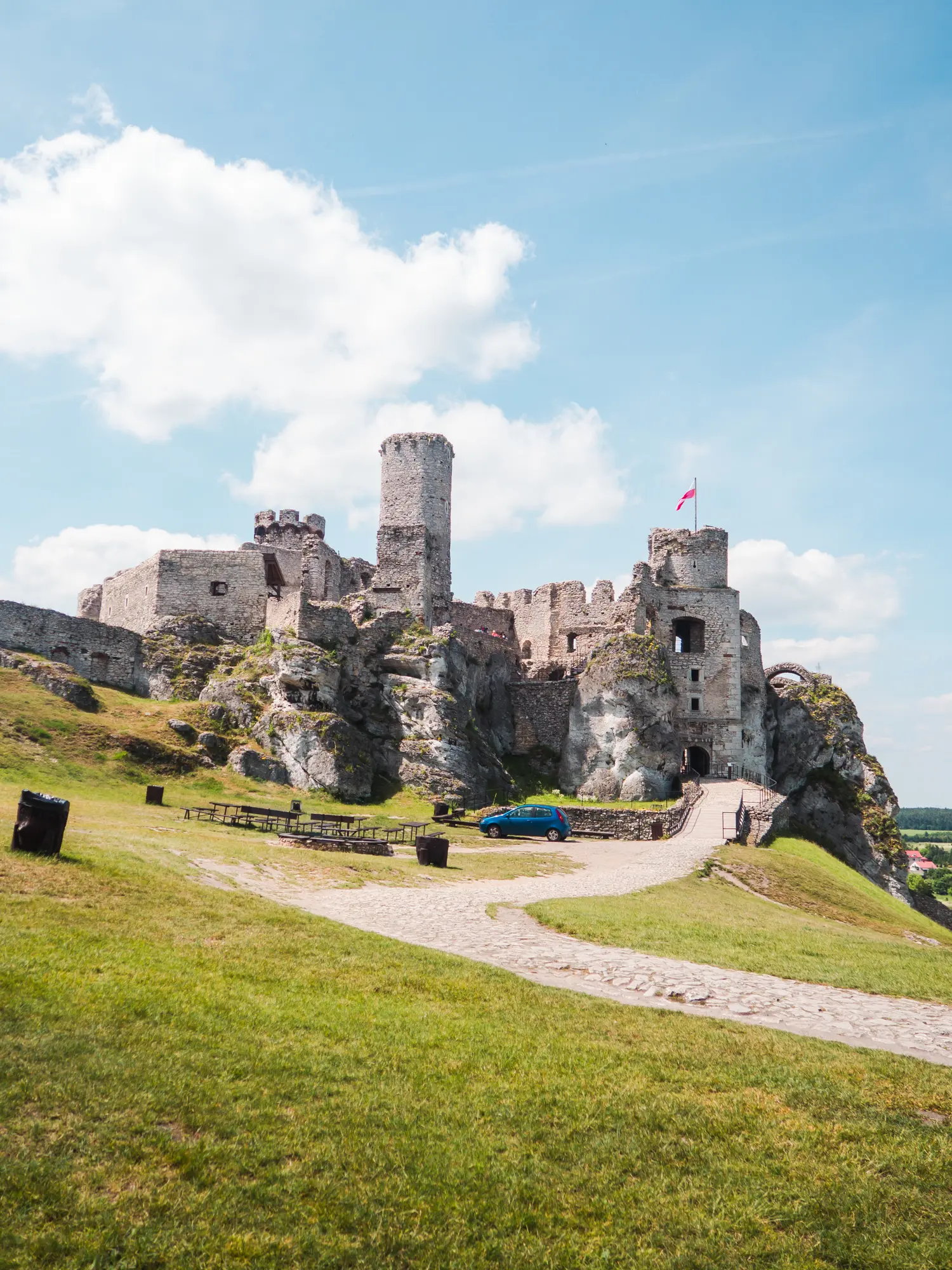 Looking towards Ogrodzieniec Castle ruin with towers and a flag, surrounded by green grass and a gravel road leading up to in on a sunny day.