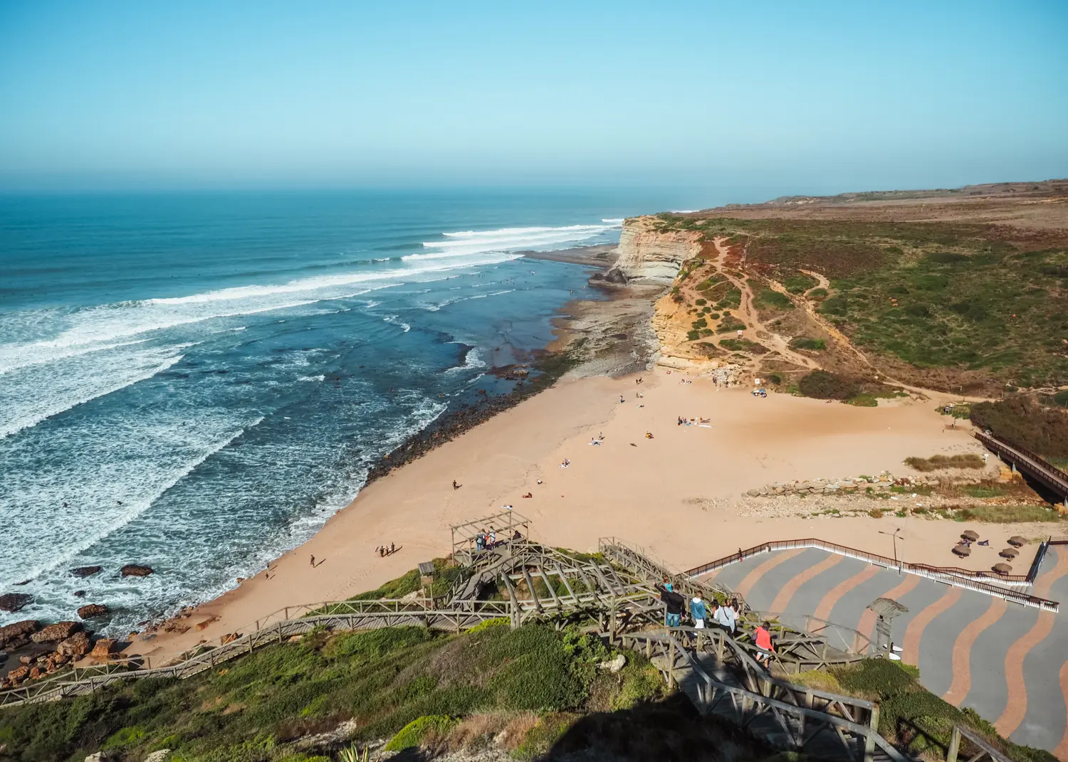 View of two headlands with a golden beach in the middle and surfers in the blue ocean on a sunny day at Ribeira d'Ilhas Beach in Ericeira.