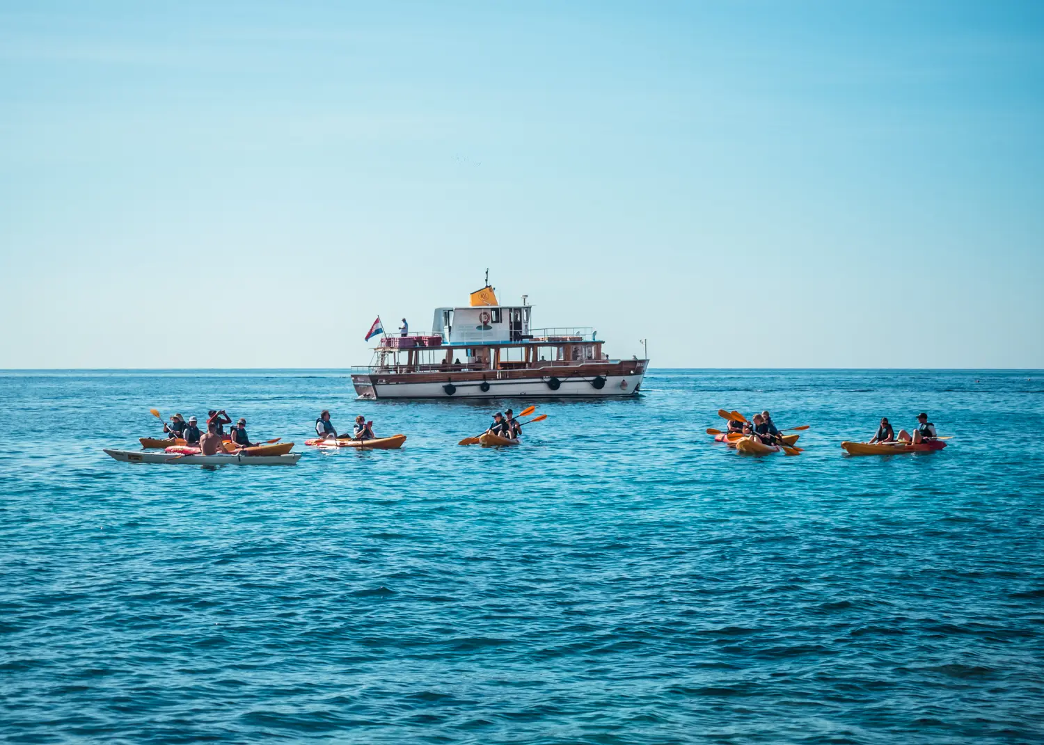 7 red and yellow double kayaks with an older tour boat in the background on the blue ocean during a kayaking tour in Dubrovnik.