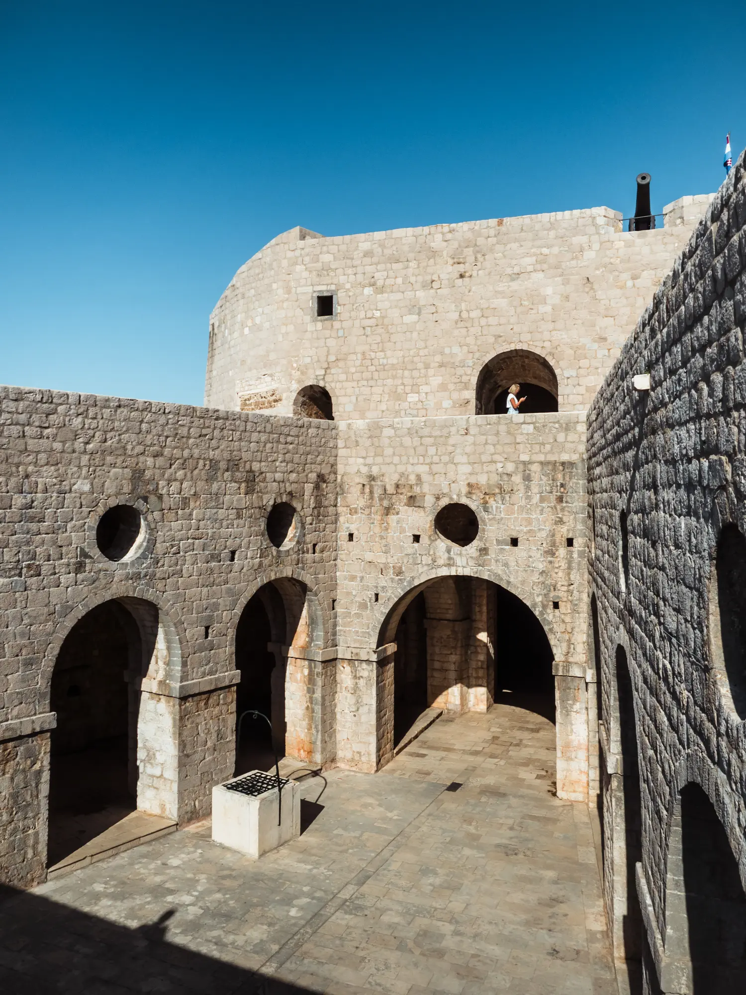 Inside stone fortress Fort Lovrijenac seen from second story looking down on inner courtyard surrounded by arches and round windows on a sunny day Game of Thrones walking tour in Dubrovnik.