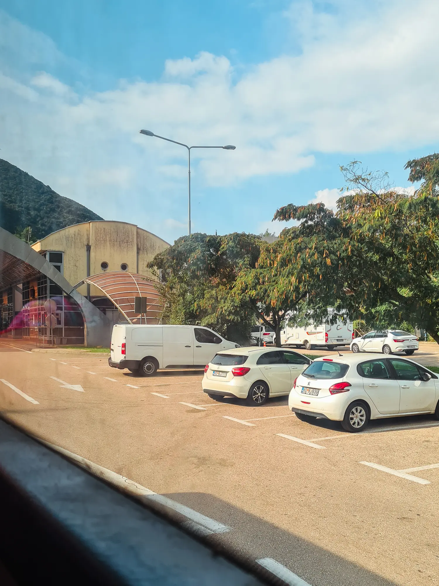 View from bus window of three white cars under a tree on a beige parking lot at the Croatia Montenegro border crossing on the way from Dubrovnik to Kotor.