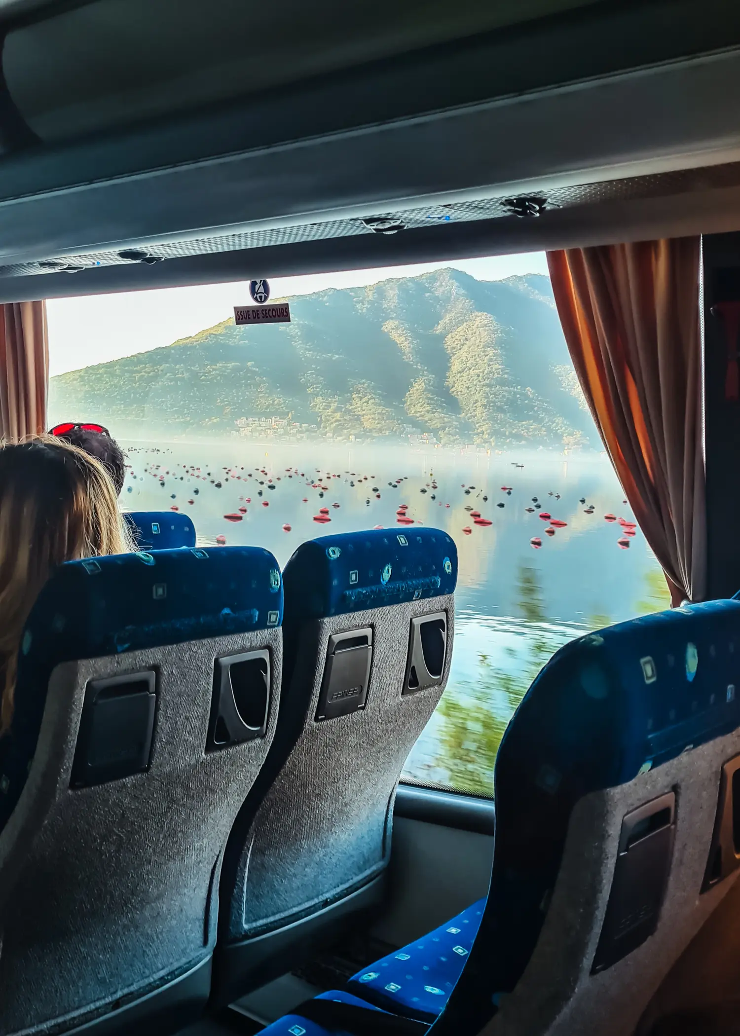 Looking out a bus window, past two blue bus seats, on the crystal clear water of Bay of Kotor with a green mountain in the background, on the way from Dubrovnik to Kotor by bus.