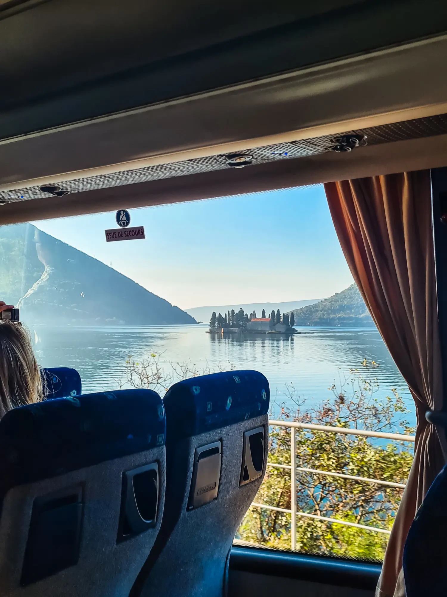 Looking out a bus window, past two blue bus seats, on a small islet in the middle of Bay of Kotor, on the way from Dubrovnik to Kotor by bus.