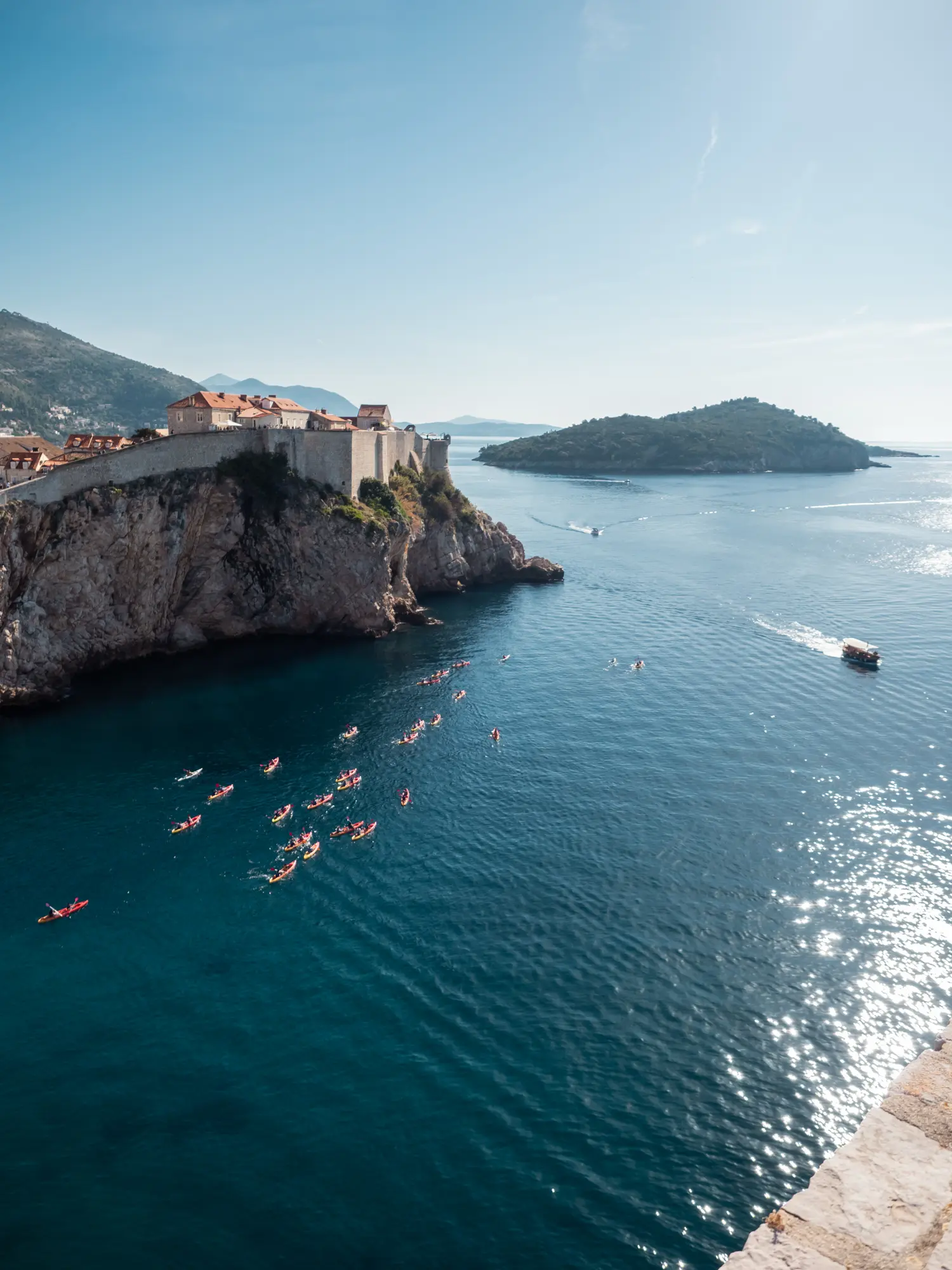 View from Fort Lovrijenac of Dubrovniks fortified Old Town and glistening water with red kayaks and two small boats in the distance during a best of Dubrovnik tour.