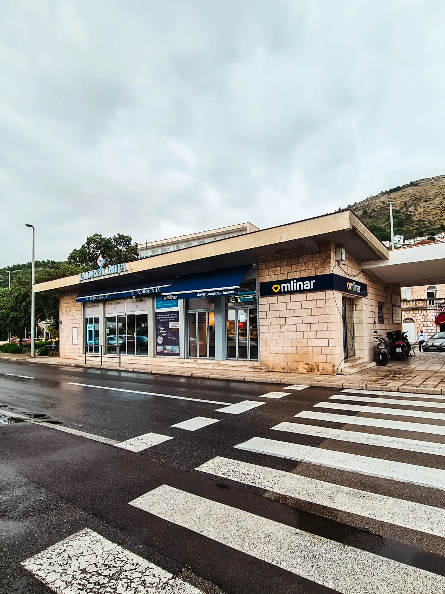 The Jadrolinija (Elaphiti Islands) Ferry Ticket office in a beige stone building by the road with a crosswalk in front in Dubrovnik on a rainy day.