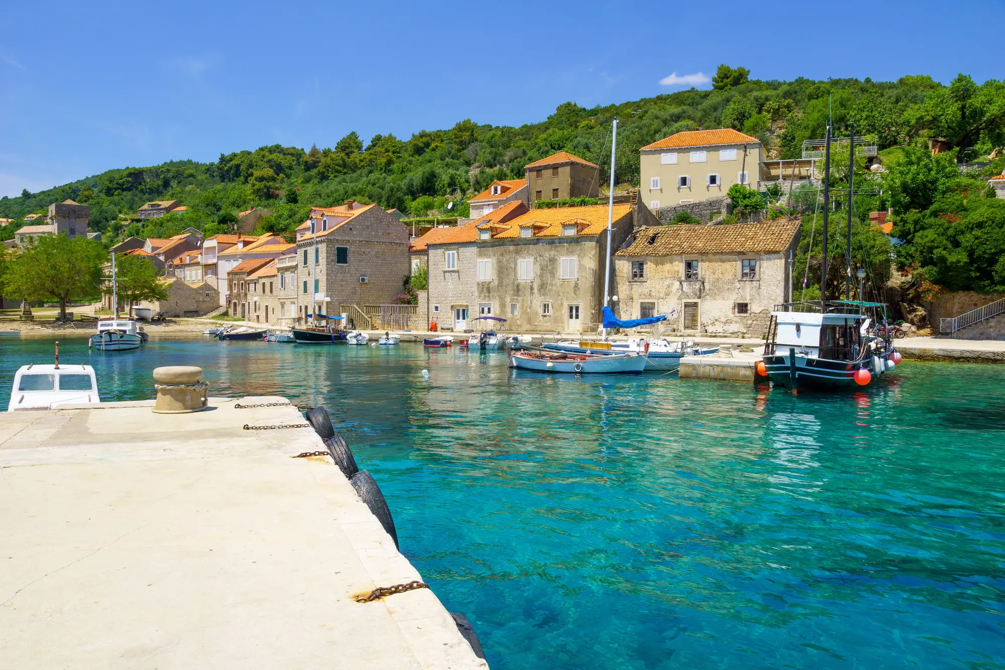 Looking towards a small harbor with boats and beige stone houses with orange roofs surrounded by green trees by the turquoise ocean in Sipan Island in the Elaphiti Islands, Dubrovnik, Croatia.