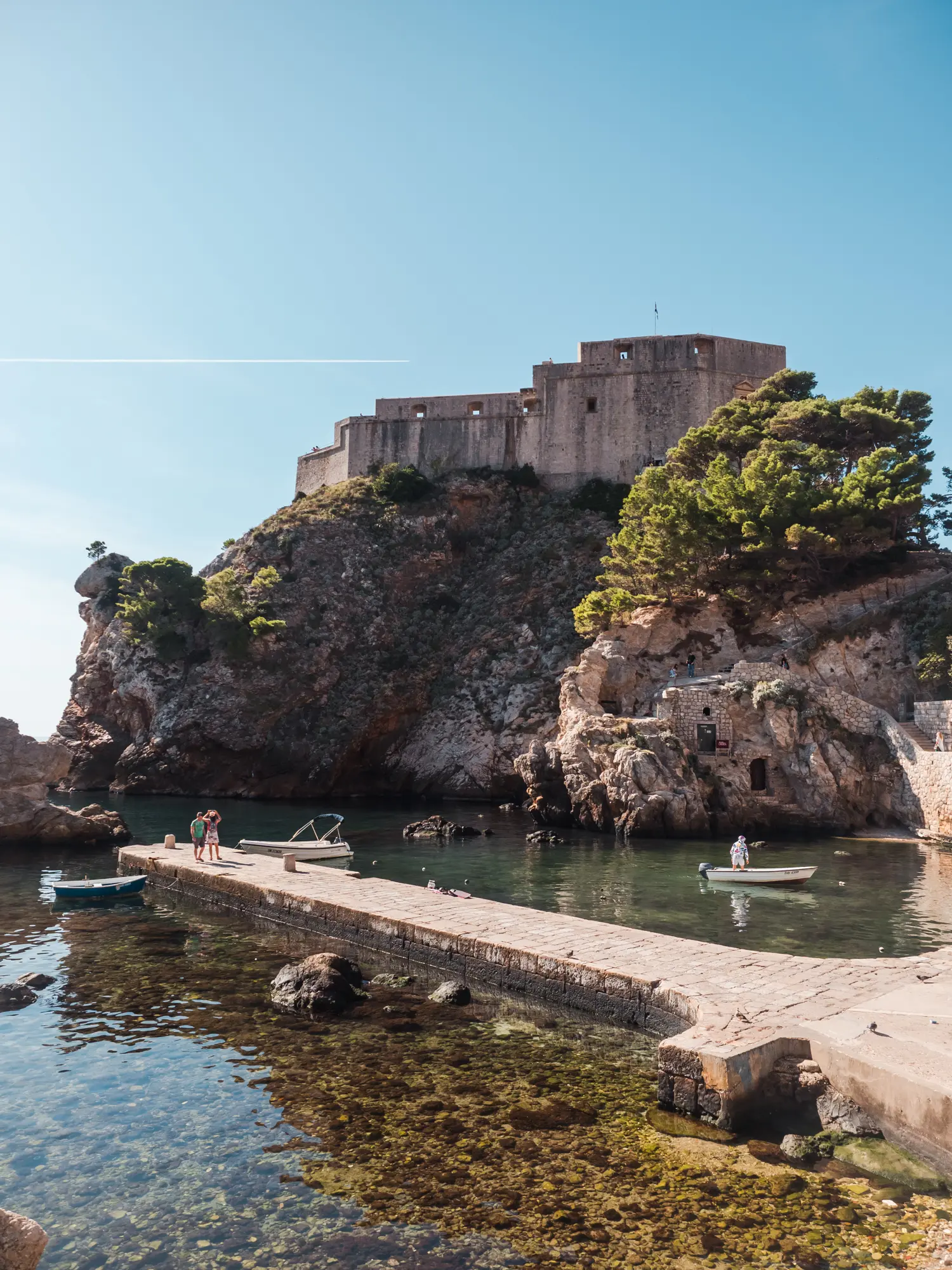 View of old stone Fort Lovrijenac on top of an outcrop behind West Harbor with a stone pier in green water during a Game of Thrones tour in Dubrovnik.