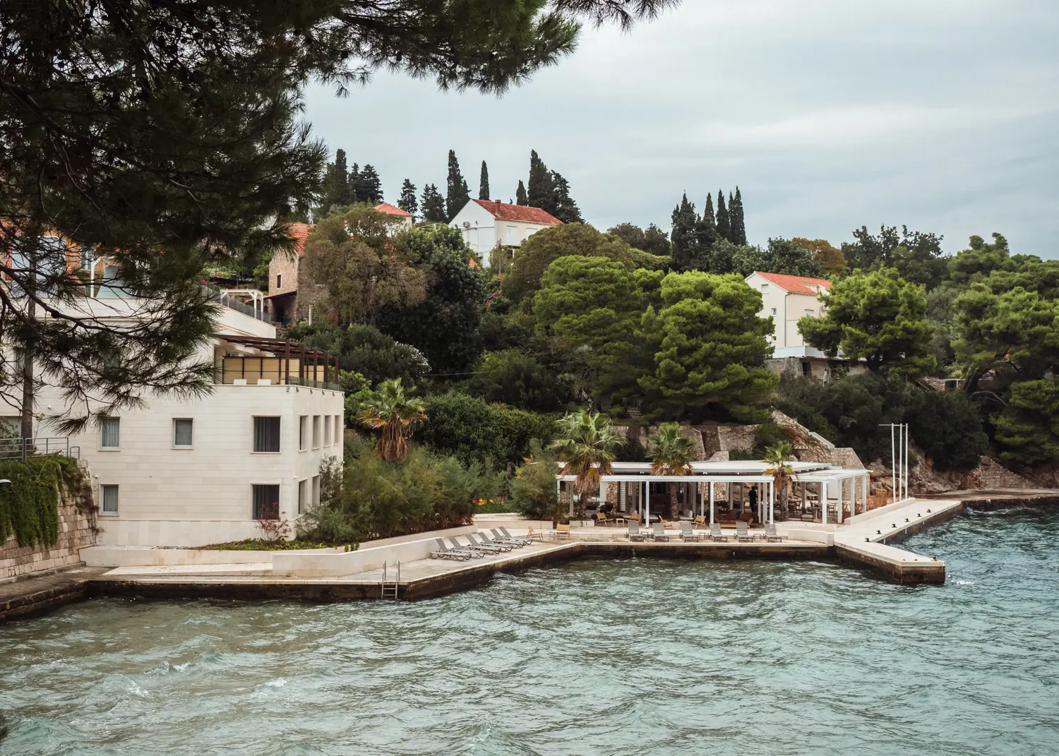 View from the ocean of the white Kalamota Beach House with a concrete lounge space and palm trees right on the ocean on Kolocep, Elaphiti Islands, Croatia.