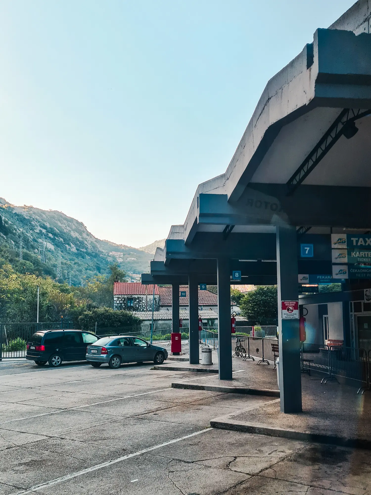 Two cars standing next to Kotor bus station with blue pillars with white wavy roof, arriving after traveling form Dubrovnik.