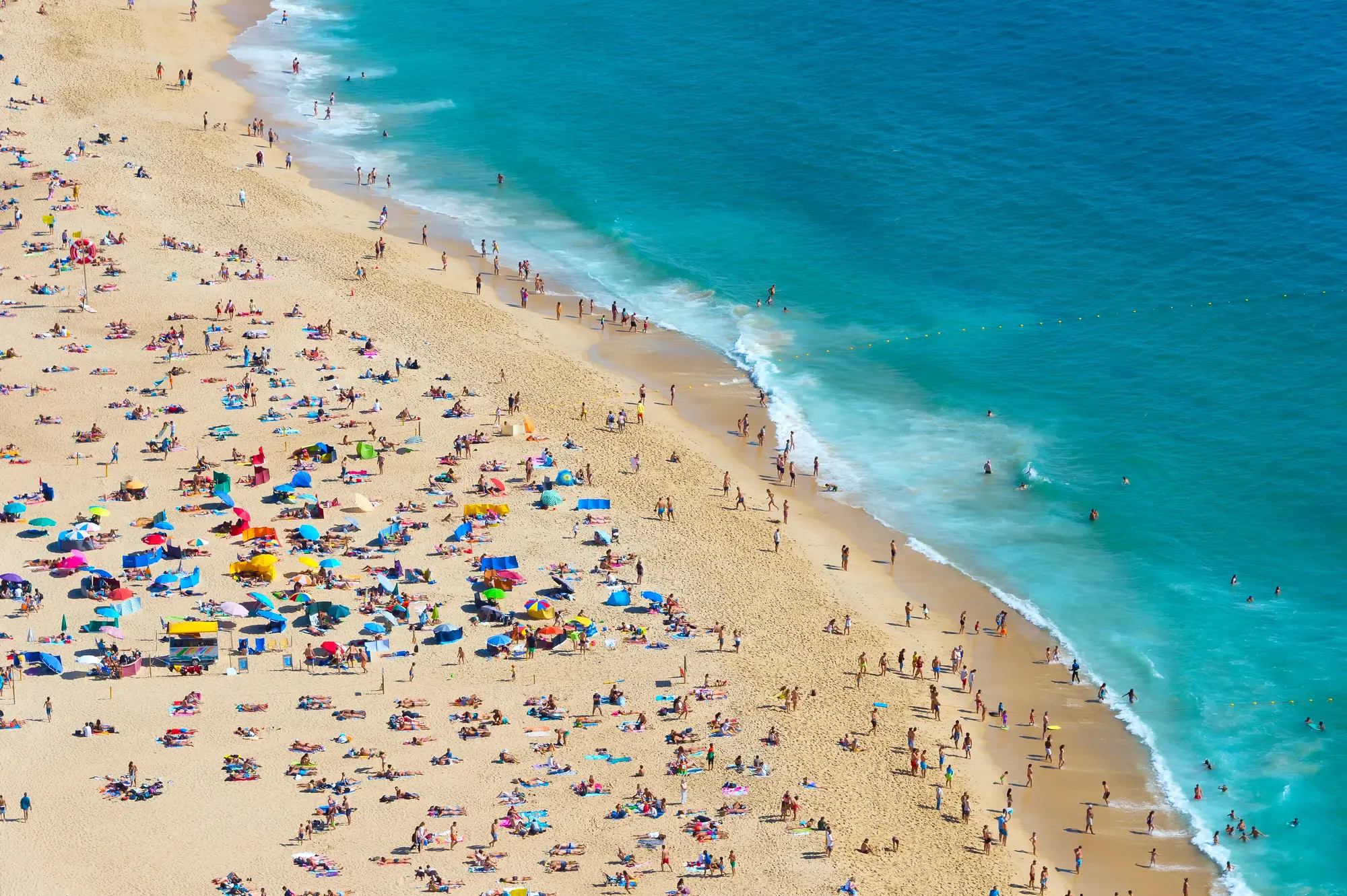 View from above of the busy Praia de Nazare beach with umbrellas in every color, lined by turquoise ocean in Nazaré, Portugal.