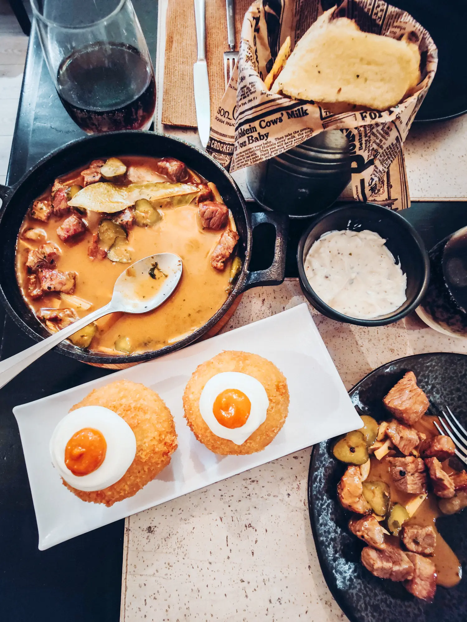 Black table with black cast iron skillets with a meat stew and a white plate with two large croquets topped with sour cream at one of the best restaurants in Nazare, Portugal.