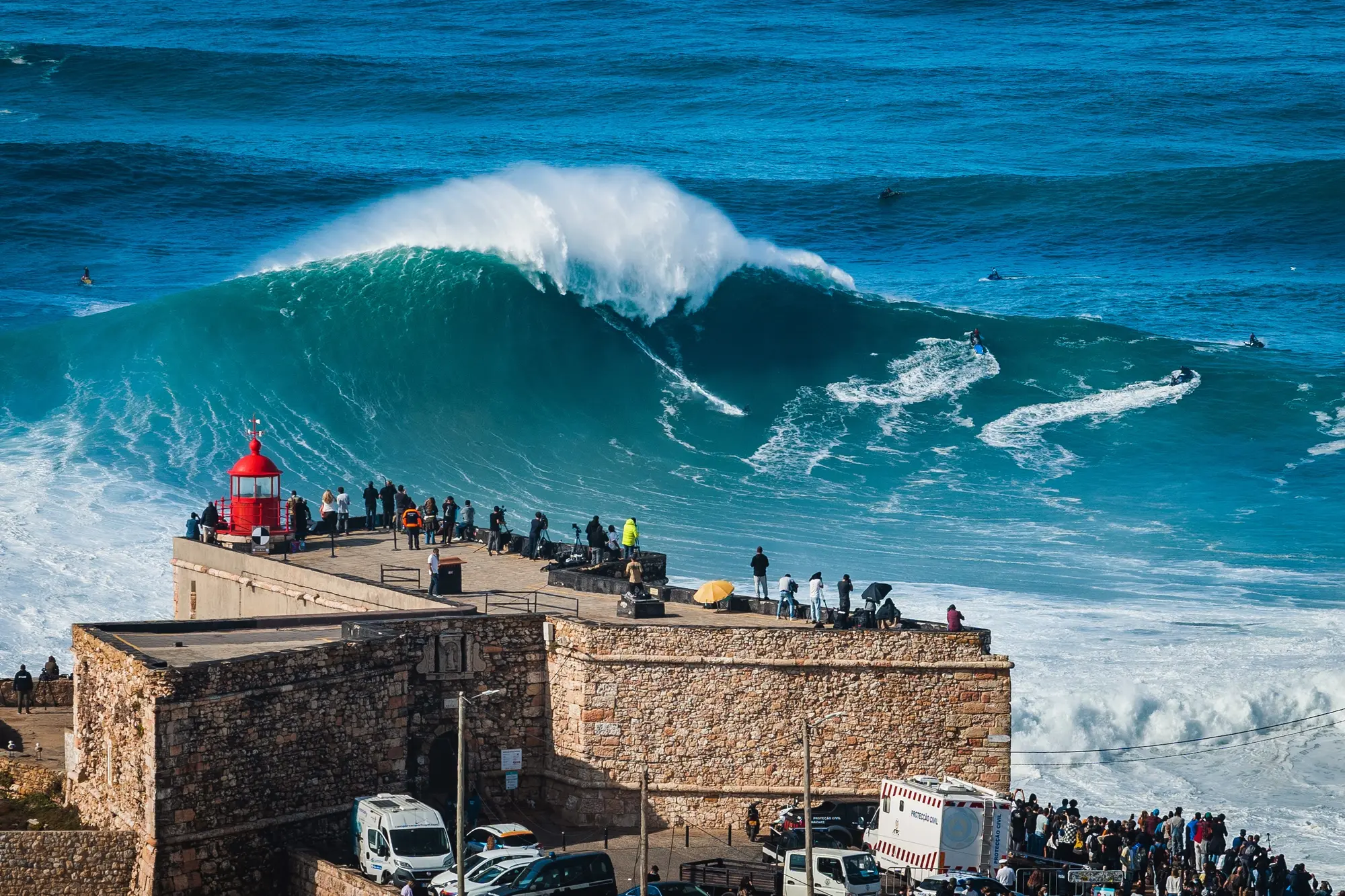People on the roof of São Miguel Arcanjo Fort watching surfers on the huge blue waves of Praia do Norte in Nazare on a sunny day.