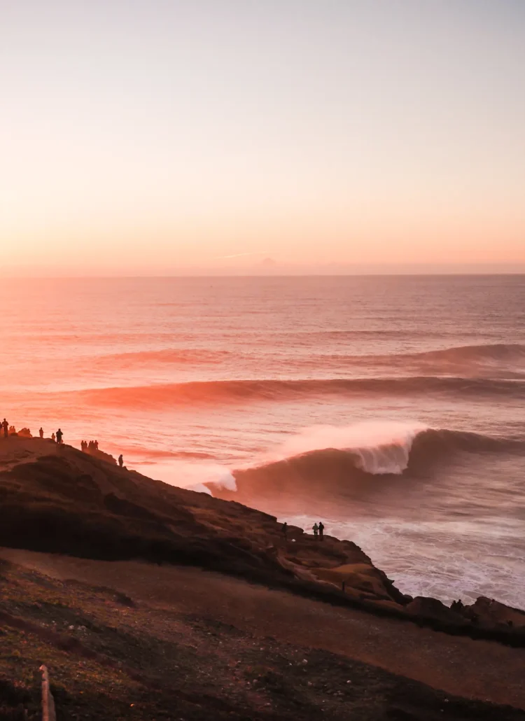 People standing on a dark cliff looking out over the huge waves at Praia do Norte during a pinkish orange sunset in Nazaré, Portugal.