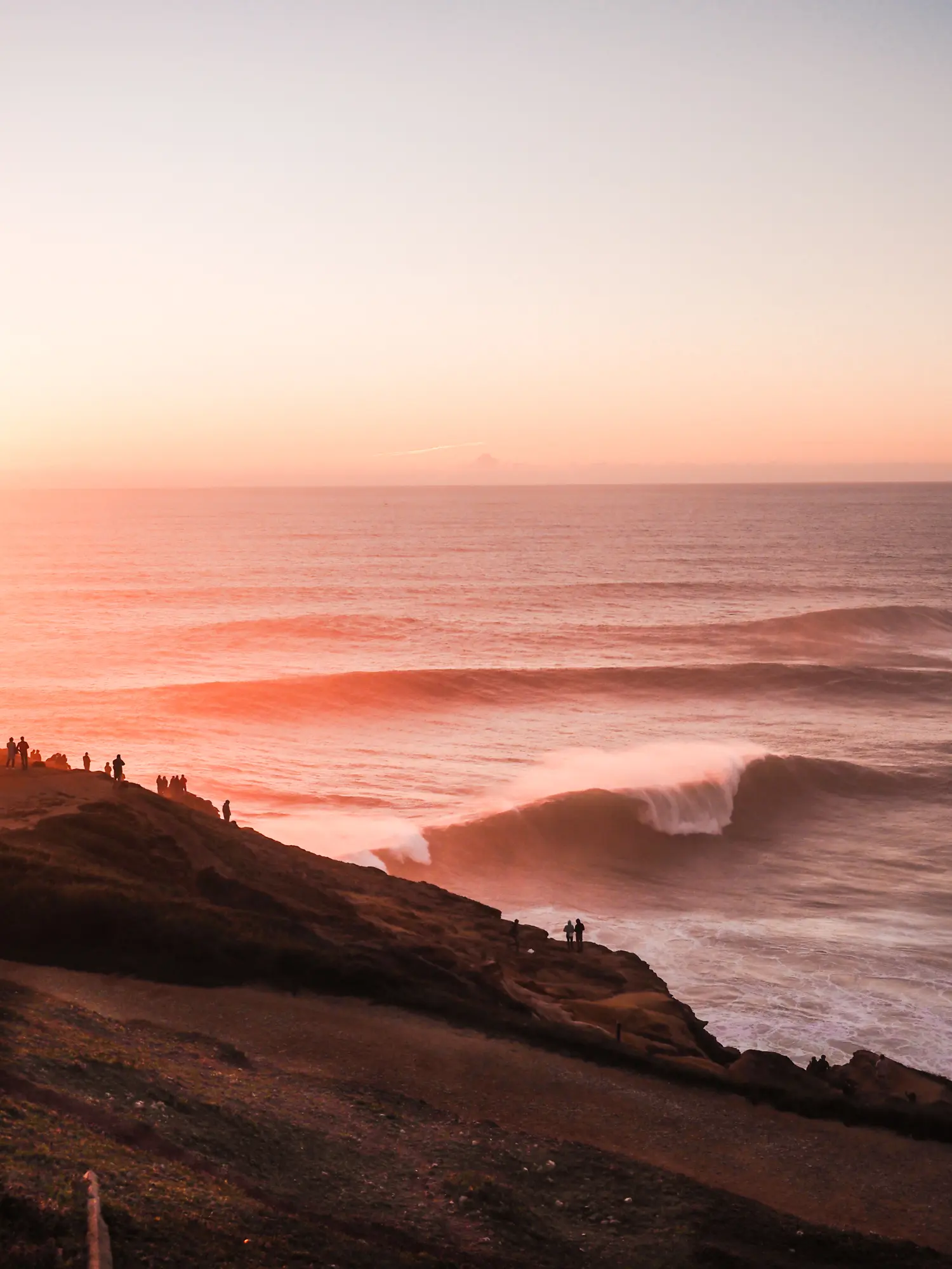 People standing on a dark cliff looking out over the huge waves at Praia do Norte during a pinkish orange sunset in Nazaré, Portugal.