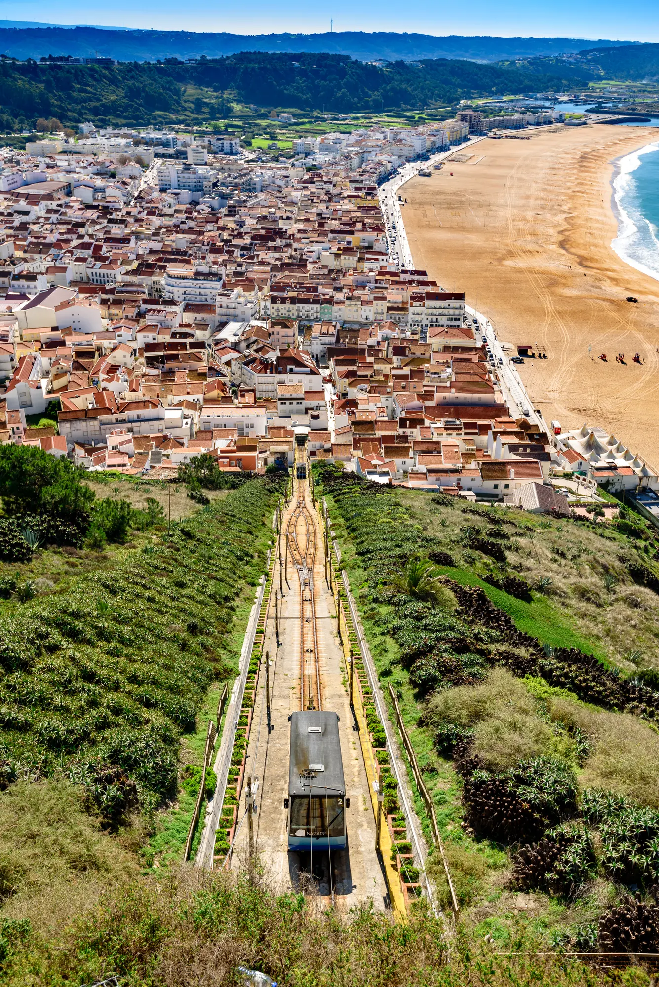 View from above of the Nazaré Funicular going down a green hillside with Praia de Nazaré beach and white houses with orange roofs in the background.