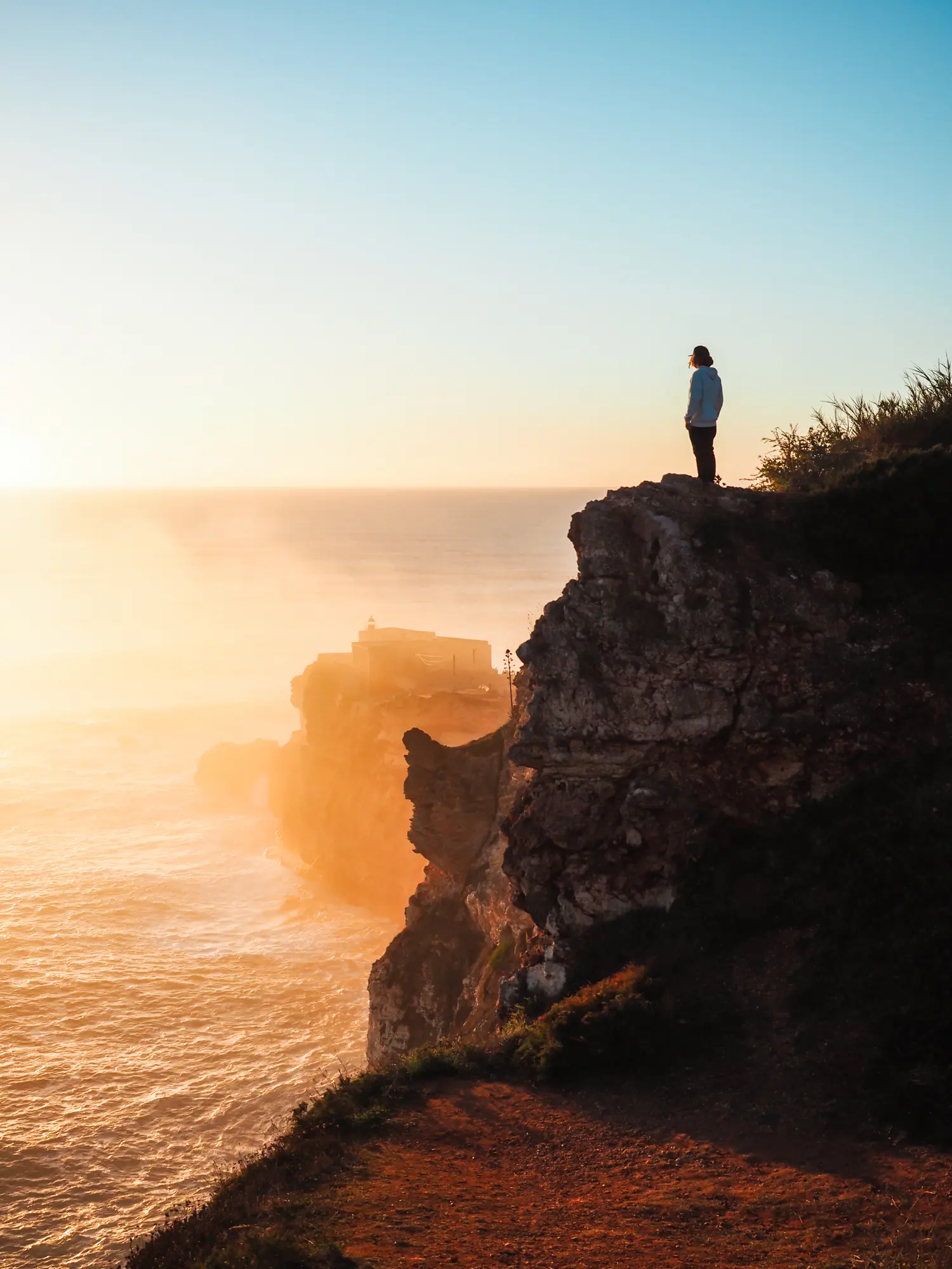 Man wearing black pant and a blue hoodie, standing atop a cliff looking out over Forte de São Miguel Arcanjo with the ocean in the background at sunset in Nazaré, Portugal.