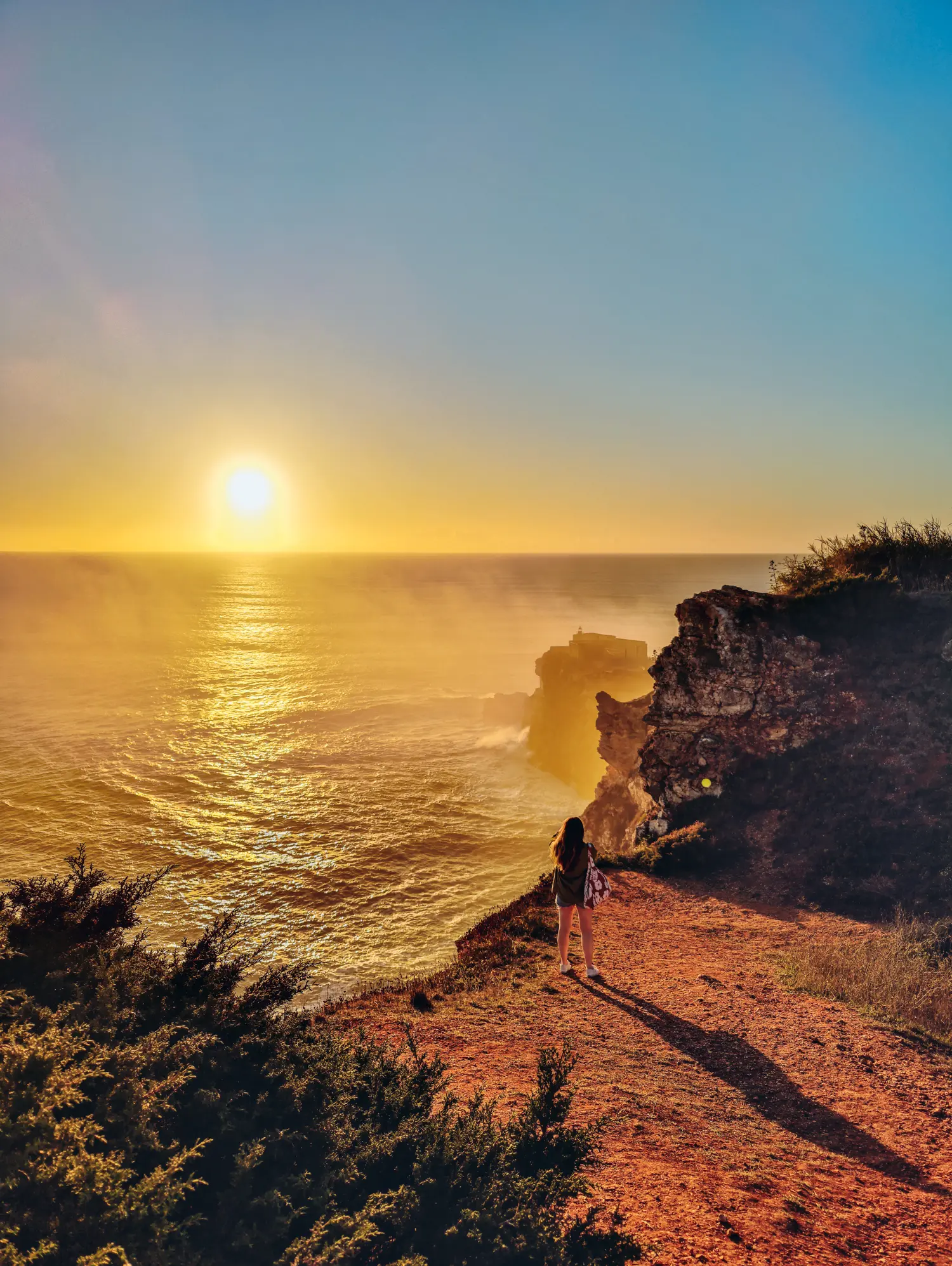 Woman standing on a viewpoint in Nazare looking out over the ocean at sunset with a fort on a rocky outcrop in the background, in Nazaré Portugal.