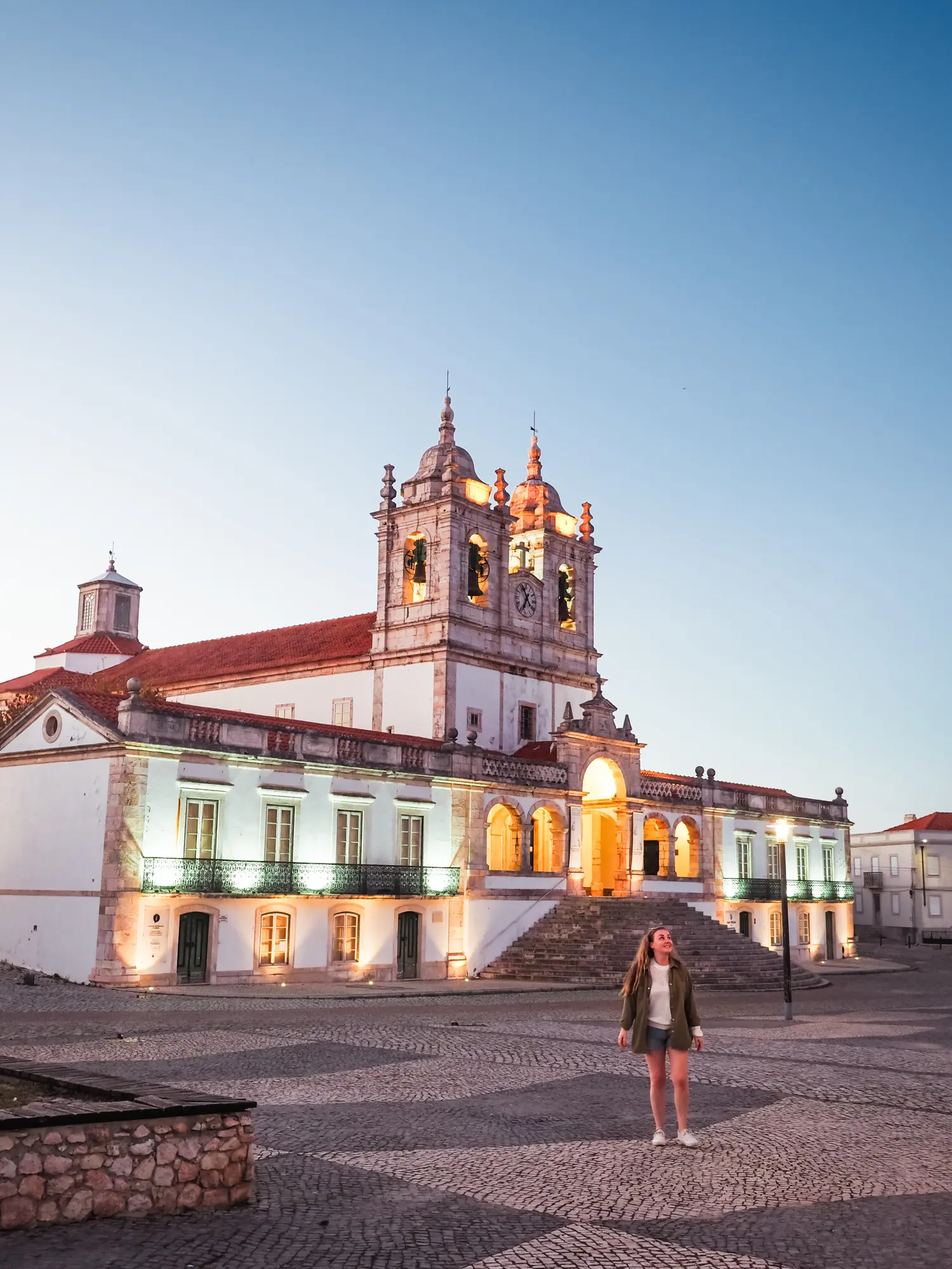 Woman wearing a green jacket, white sweater and denim shorts, standing in a bog cobbled square in from of the lit up Sanctuary of Our Lady of Nazaré, a beautiful old building in Nazaré, Portugal at dusk.