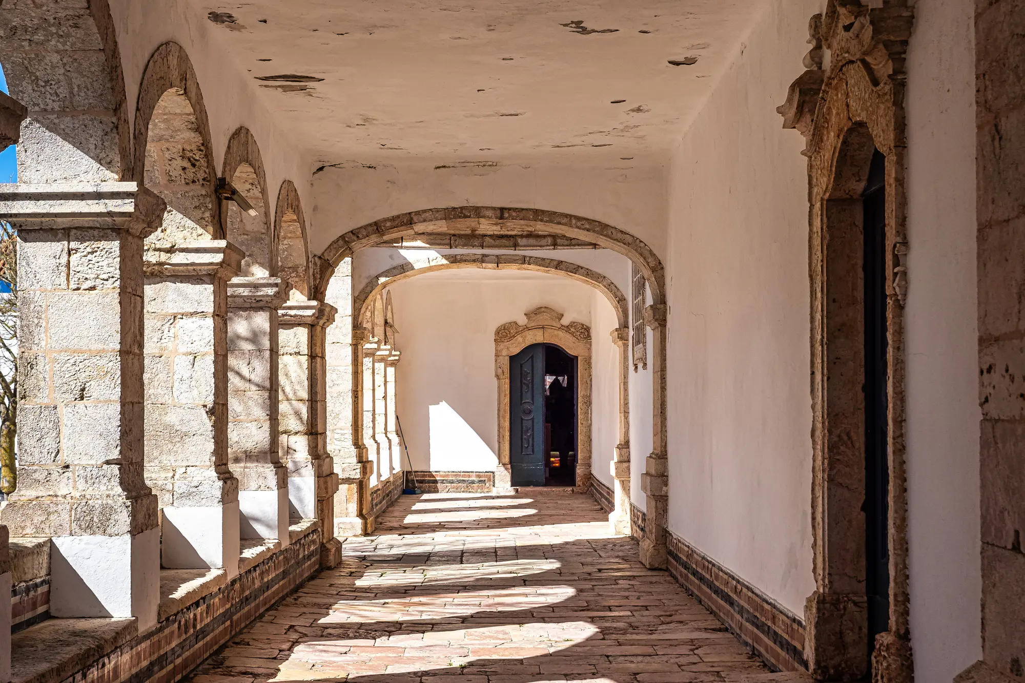 Sun shining in between stone arches in a covered walkway in an old white church in Nazaré Portugal.